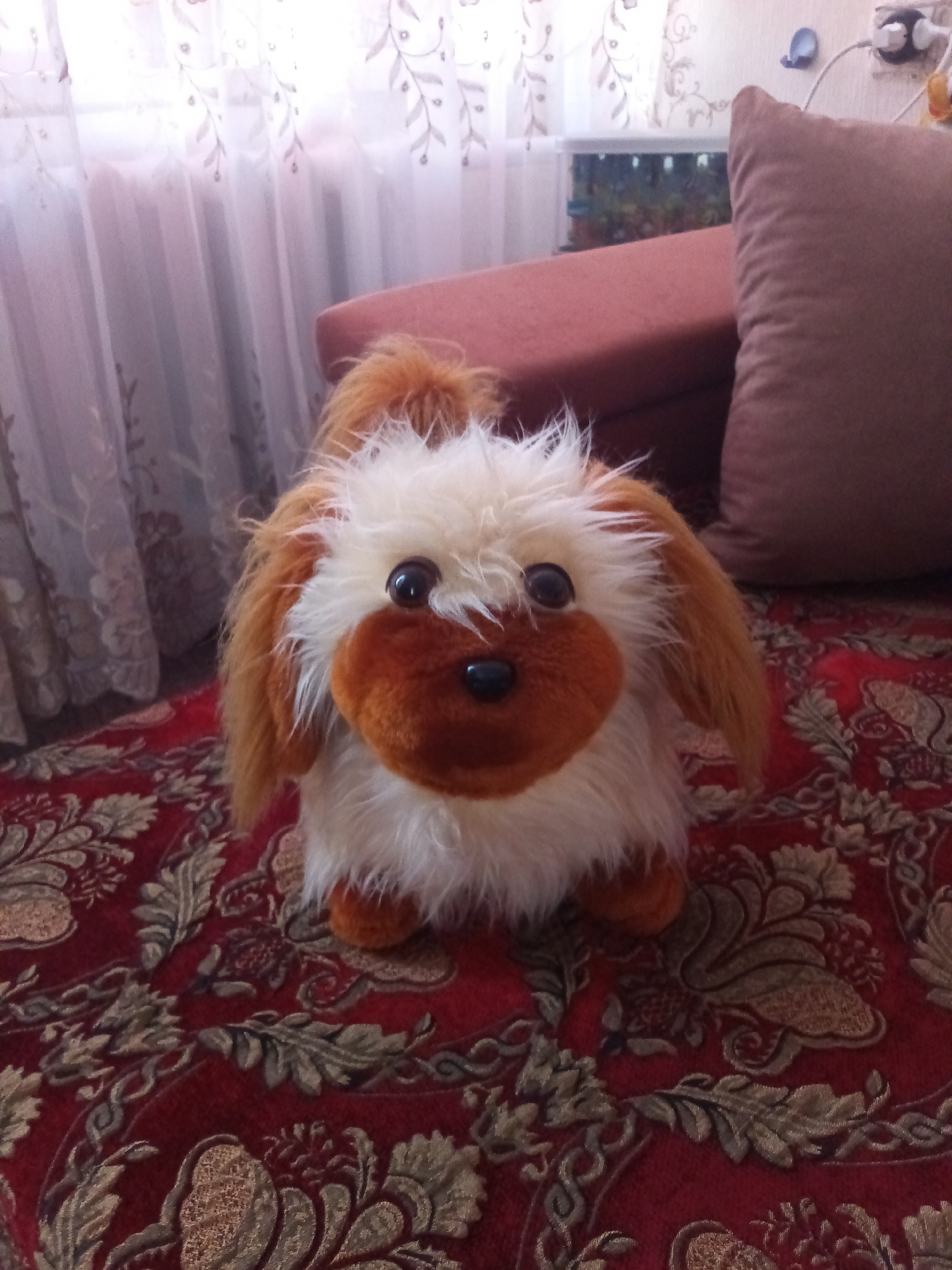 A plush toy dog with fluffy white and tan fur sits on a red, ornate rug. The dog has big, dark eyes and a brown snout. Its ears are long and tan. The background is blurred but appears to be an indoor setting. To the left, sheer curtains with a floral pattern hang, suggesting natural light from a window. Behind the dog, a plush brown cushion and a reddish-brown piece of furniture are visible. In the distance, an aquarium with lights can be seen. The scene is well-lit, likely during the daytime. There is no visible text in the image. The context provided indicates the location is Chernyshevskoye, Russia.