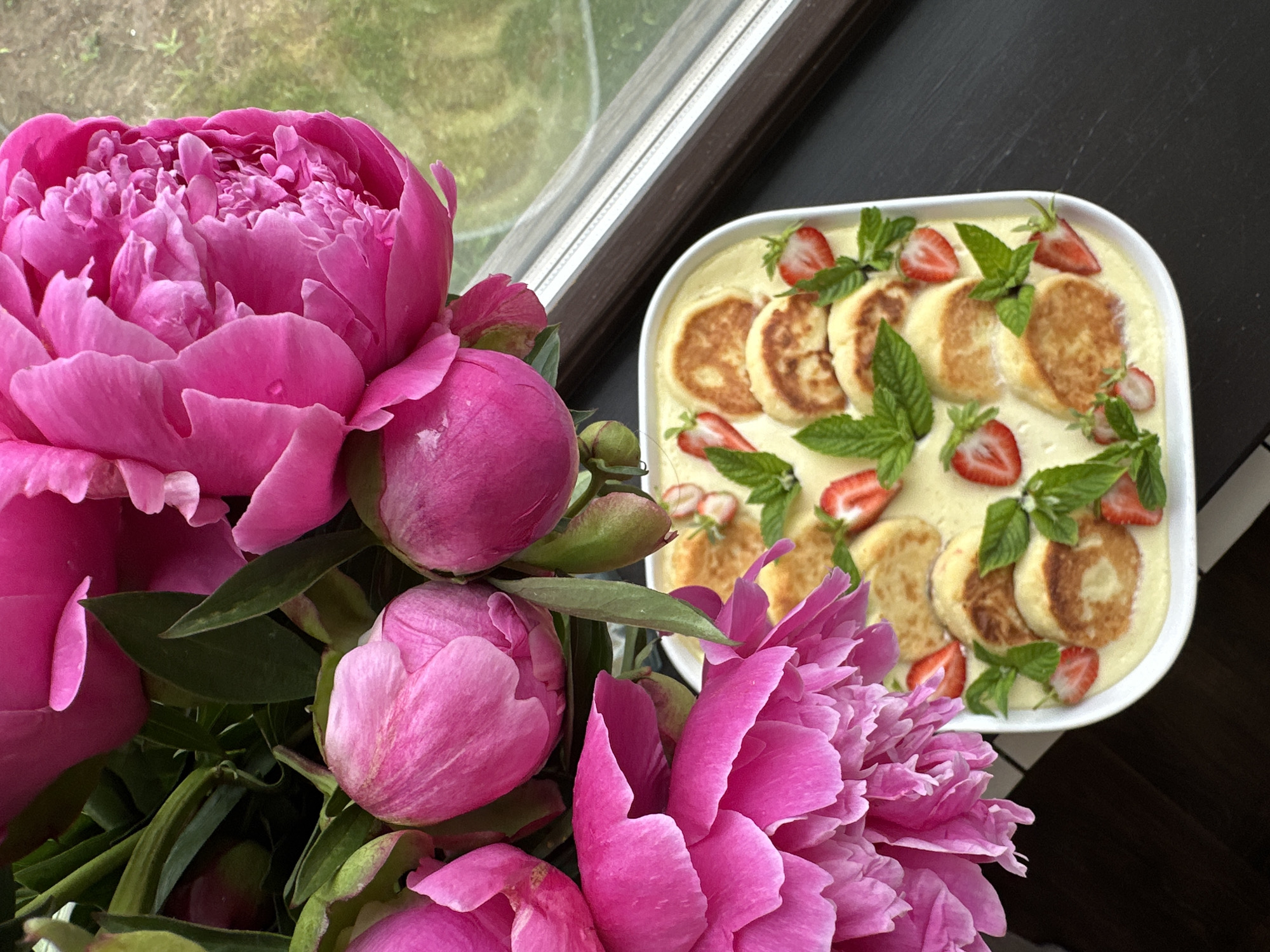 A close-up, top-down shot captures a white ceramic dish filled with small, golden-brown pancakes in a creamy sauce. The pancakes are artfully arranged and garnished with fresh mint leaves and sliced strawberries. To the left, a lush bouquet of vibrant pink peonies dominates the frame, their petals unfurling in various stages of bloom. The background features a dark surface, possibly a table, and a window with a blurred green outdoor scene visible through it. The overall impression is one of a beautifully presented dessert, set against a backdrop of floral elegance, likely in a domestic setting in Chaikovsky, Russia, given the context.