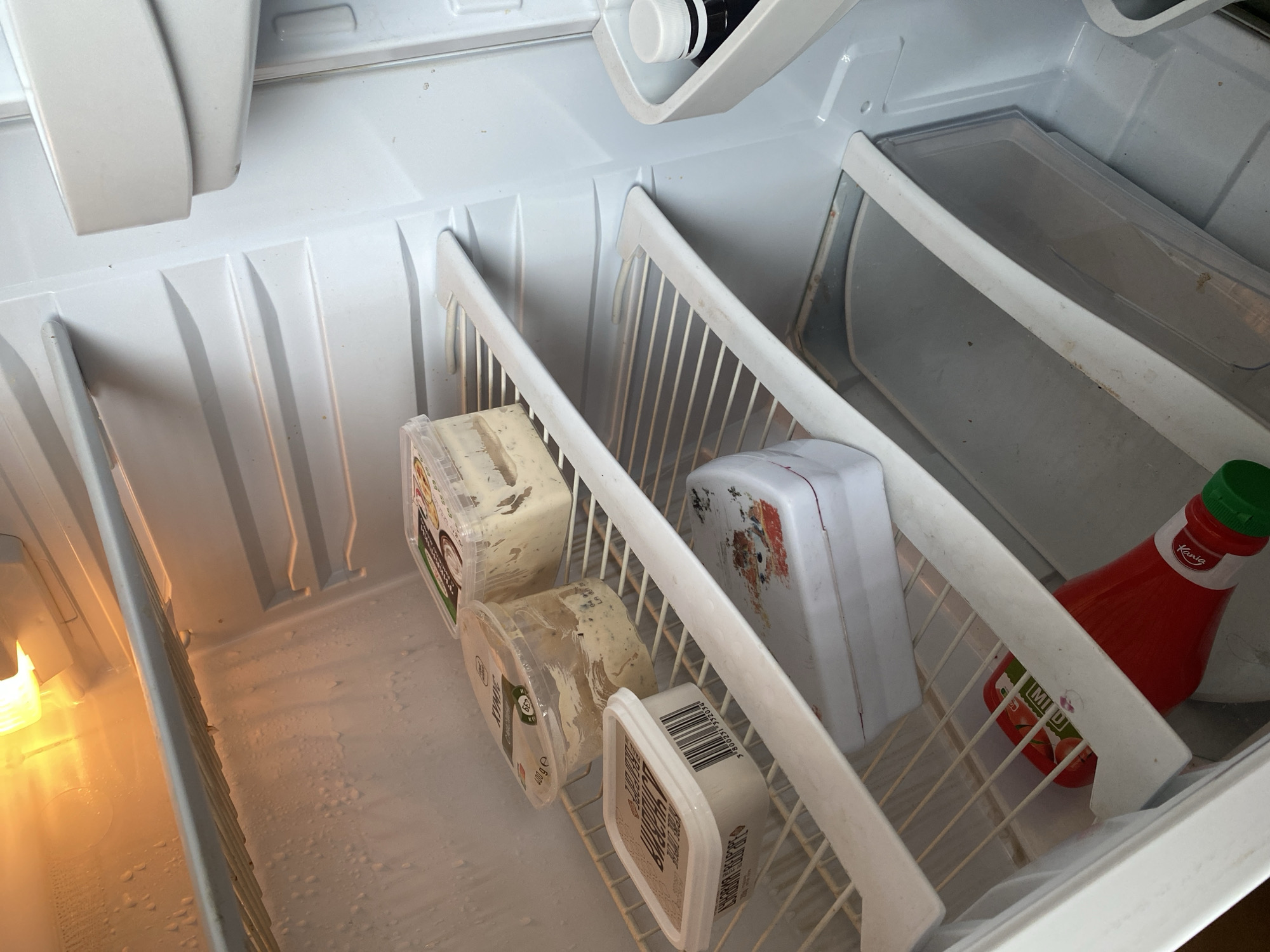 The interior of a refrigerator is shown. The refrigerator interior is white. There is food in the fridge, including containers of what appears to be spreadable cheese or dips, a white floral box, and a bottle of ketchup. The shelving is white. The interior surfaces of the refrigerator show water droplets and some smudges or food residue. There is an interior light on. The image was taken in Sofia, Bulgaria.