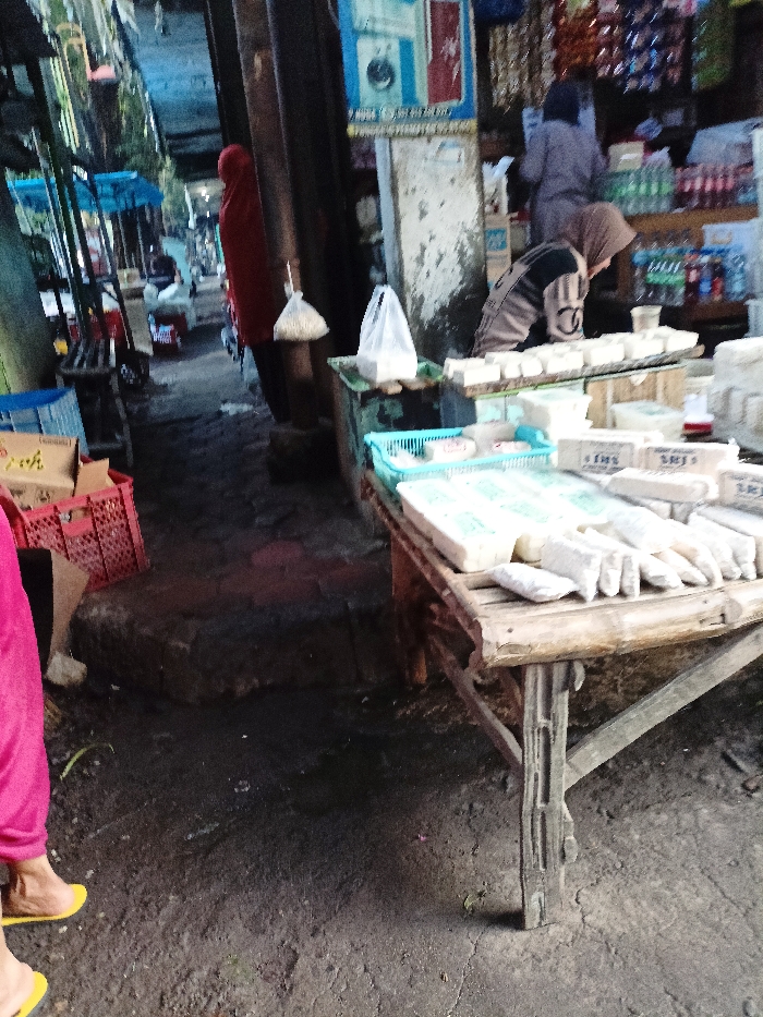 This image captures a vibrant market scene in Mojosari, Indonesia, set within a semi-enclosed or covered alleyway. The ground is a dark, unpaved surface, suggesting a traditional outdoor market environment.
In the foreground on the left, a person wearing bright pink/red pants and yellow sandals is partially visible, indicating movement or standing near the path. In the mid-ground left, another individual dressed in red clothing and a matching head covering stands partially obscured by a dark column.
The right side of the image features two market stalls, constructed from simple wood and bamboo. The front stall displays numerous white blocks, likely tofu or tempeh, some individually wrapped. A light blue plastic basket sits on this stall, along with plastic bags, one of which contains small white granular items. A person wearing a light brown head covering leans over this stall, seemingly engaged in selling or arranging products. Further back, behind the stalls, another person in grey clothing is partially visible near shelves stocked with various bottled drinks and packaged goods.
A poster is affixed to a column in the background, featuring an image of what appears to be a washing machine or similar appliance, alongside red sections and indistinct text. Faint text, possibly "FJS" and "LE," can be seen on some of the white food packaging on the stalls. The natural lighting suggests it is daytime, with shadows indicating the sun is not directly overhead. The scene depicts typical daily activity within a local market.