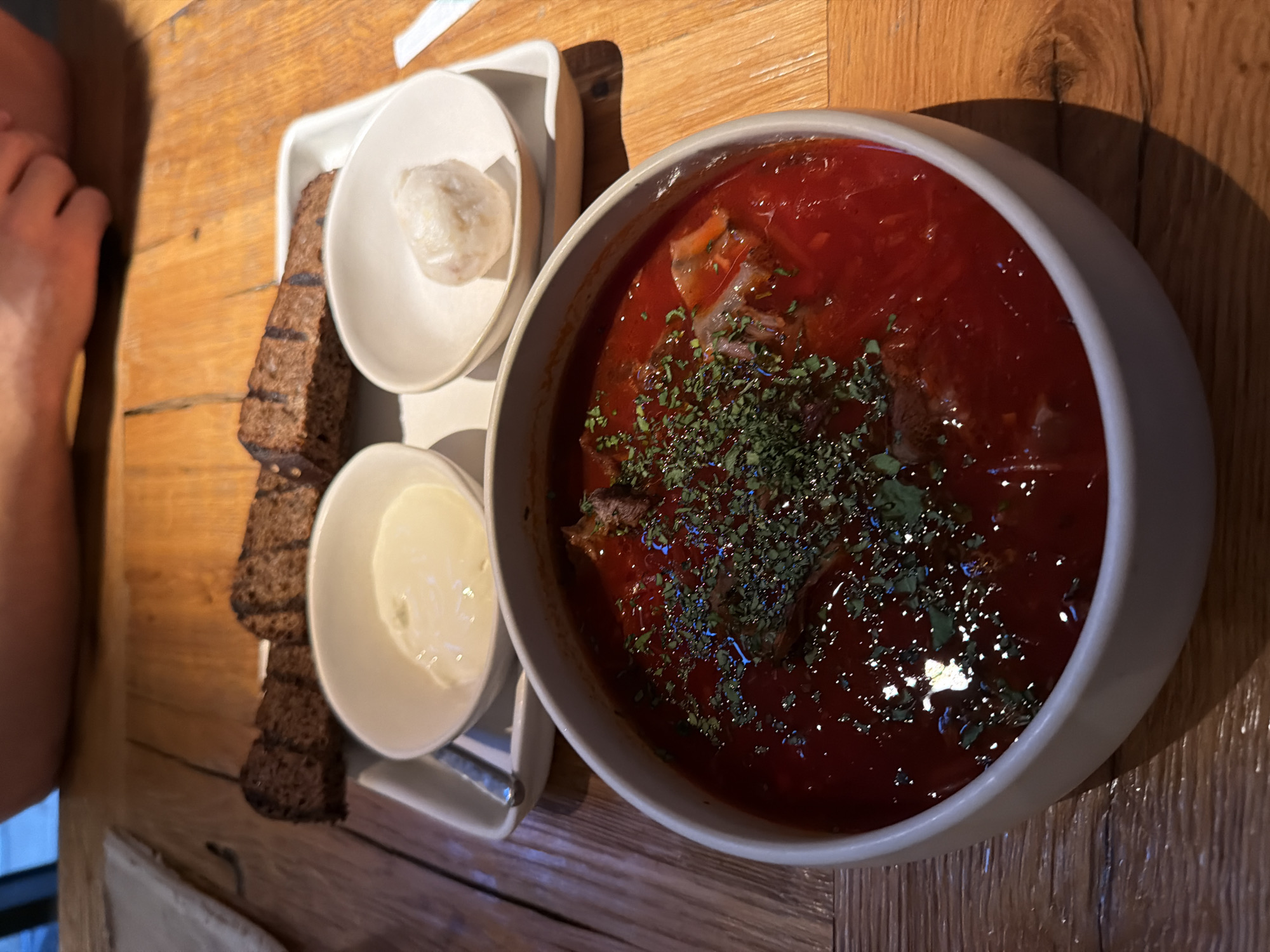 A high-angle shot depicts a bowl of borscht, a traditional Eastern European soup, garnished with fresh herbs, set on a wooden surface. Beside the bowl, on a small white tray, are slices of dark bread and small bowls containing what appears to be sour cream and possibly garlic paste. The scene is brightly lit with natural light, creating distinct shadows on the table surface. The composition suggests the image was taken in an indoor dining setting, likely a restaurant or cafe. The location in Moscow, Russia, is inferred due to borscht's prevalence in Russian cuisine.