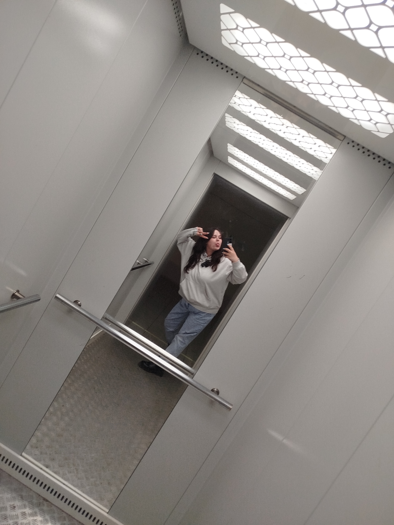 A young woman with long, dark hair takes a selfie in an elevator. She wears a light-colored hoodie and blue jeans and sticks her tongue out while holding up a peace sign. The elevator has silver metal walls and a non-slip metal floor. The ceiling has a patterned light fixture. The location is unknown and cannot be determined from the image. The city of Chaikovsky, Russia cannot be confirmed as the location.