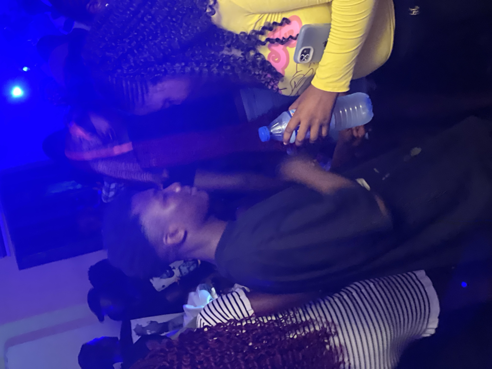 The image depicts a dimly lit indoor scene, possibly a nightclub or party, with predominantly blue and purple lighting. In the foreground, a young man with short, dark hair is seen from the side, wearing a dark t-shirt. To his right, a young woman with long, curly dark hair is visible, wearing a bright yellow long-sleeved top with a cartoonish design and black pants. She is holding a clear plastic bottle of water in her right hand and a light gray smartphone in her left hand, with the phone's camera visible. Another person with long, dark curly hair and a striped t-shirt is partially visible on the left side of the frame. Other individuals are present in the background, largely obscured by the low lighting and the presence of other people. The overall atmosphere suggests a social gathering. The location is identified as Wumat, Nigeria.