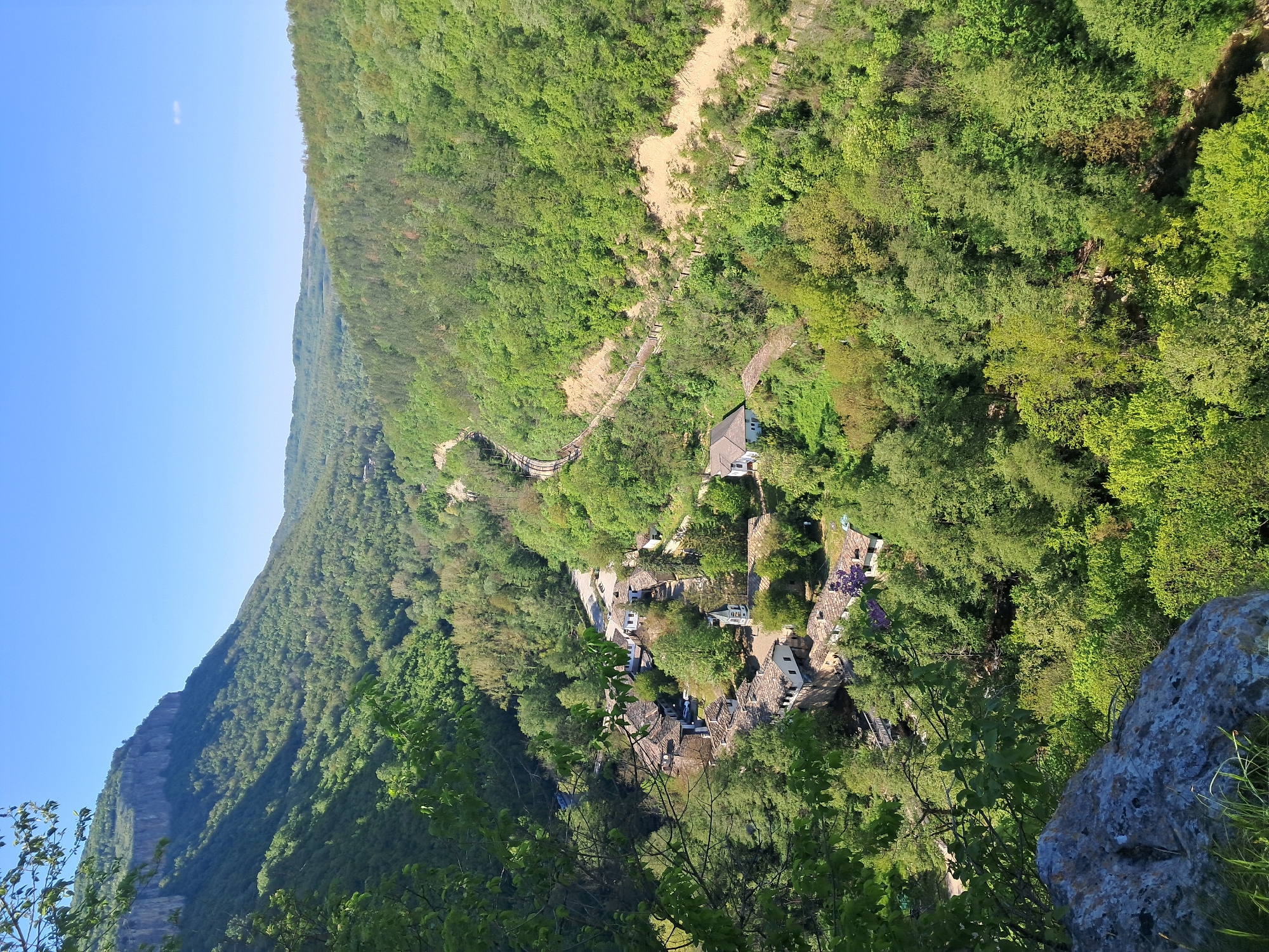 The image captures a view looking down into a valley near the city of Dryanovo, Bulgaria. The landscape is dominated by lush green trees covering the slopes of the surrounding hills. A small settlement or village is nestled in the valley floor, featuring buildings with dark roofs, possibly made of stone or tile, interspersed with trees. A winding dirt path or road is visible leading up one of the hillsides, seemingly connecting the village to higher elevations. In the distance, a sheer rock face stands out prominently on one side of the valley. The sky is a clear blue. The vantage point appears to be from a high elevation, possibly a rocky outcrop or cliff, overlooking the scene.