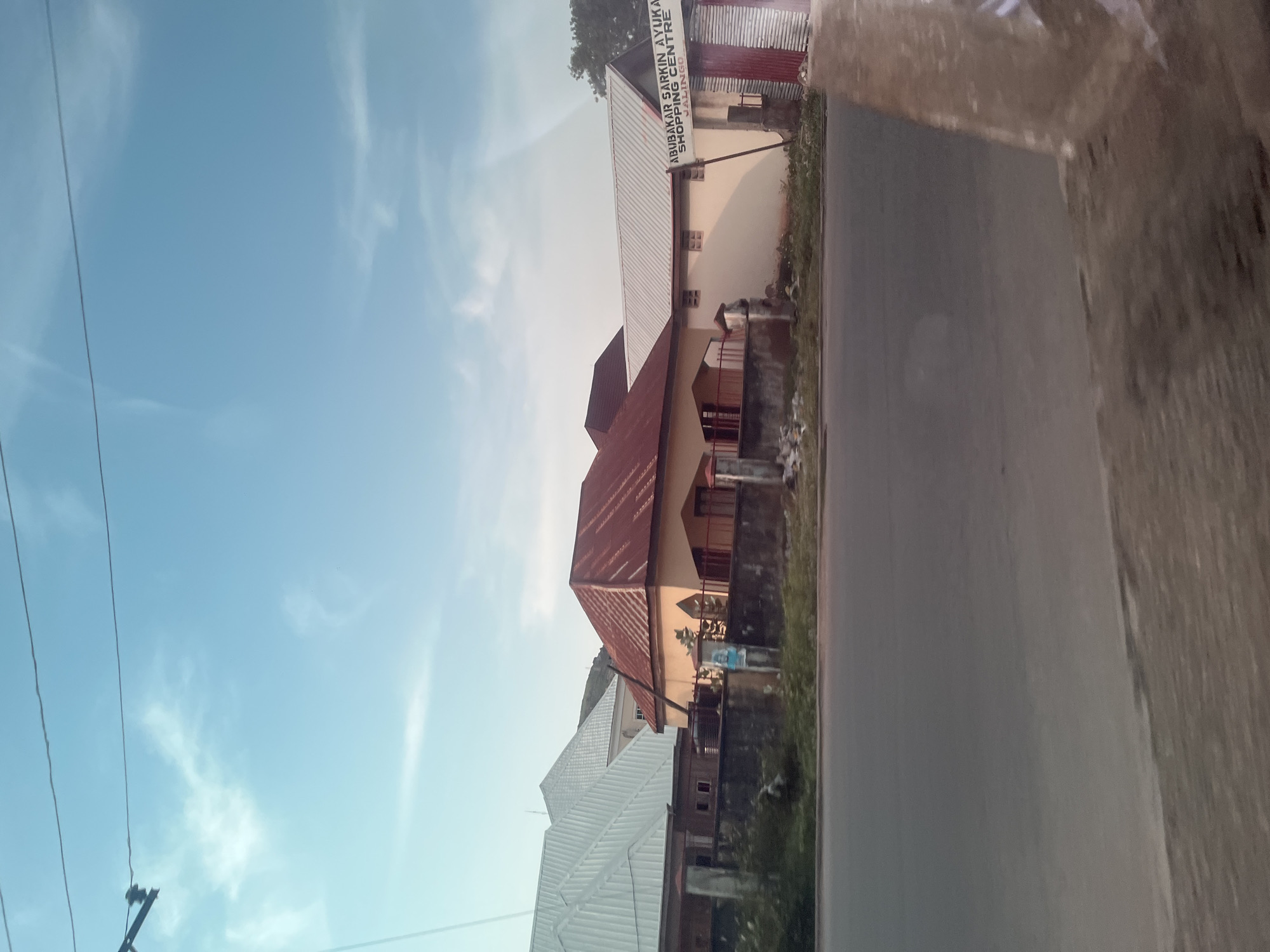 The image shows a street scene in Jalingo, Nigeria, with buildings and a road. The photograph is oriented vertically, with the sky at the left and the road at the right. There is a building with a red metal roof and a lighter colored facade. Above this building, a white sign is visible with red lettering. The text on the sign reads "ABUBAKAR SARKI AYUKAN SHOPPING CENTRE JALINGO". Another building with a white metal roof is visible behind the first building. The sky is clear with some wispy clouds. The time of day appears to be late afternoon or early evening, as indicated by the lighting and the blue hue of the sky. The weather seems fair. The scene depicts a typical street in a Nigerian town, with a mix of commercial and residential buildings. There are no people or specific activities clearly visible in the image. The overall impression is one of everyday urban life.