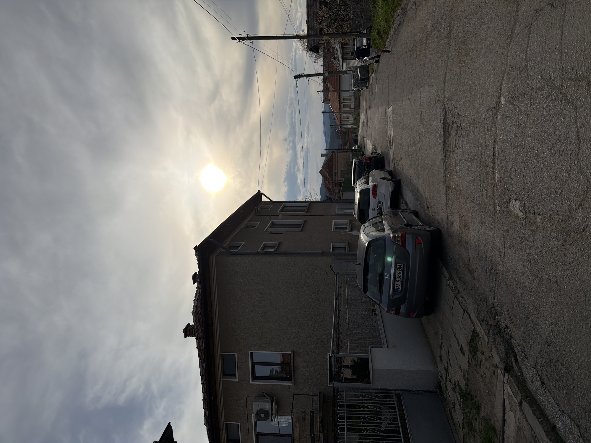 The image depicts a residential street in Popintsi, Bulgaria. A three-story building occupies the left side of the frame, and several parked cars are visible on the right side of the street. The sky is partly cloudy with the sun shining through. Power lines are seen stretching across the top of the image, and distant mountains are visible in the background. The street is asphalt, showing cracks and signs of wear. The front of the building has an air conditioning unit. A pedestrian is walking in the street near the top of the frame. The car closest to the camera has a Bulgarian license plate that reads "PA 8386 BM".