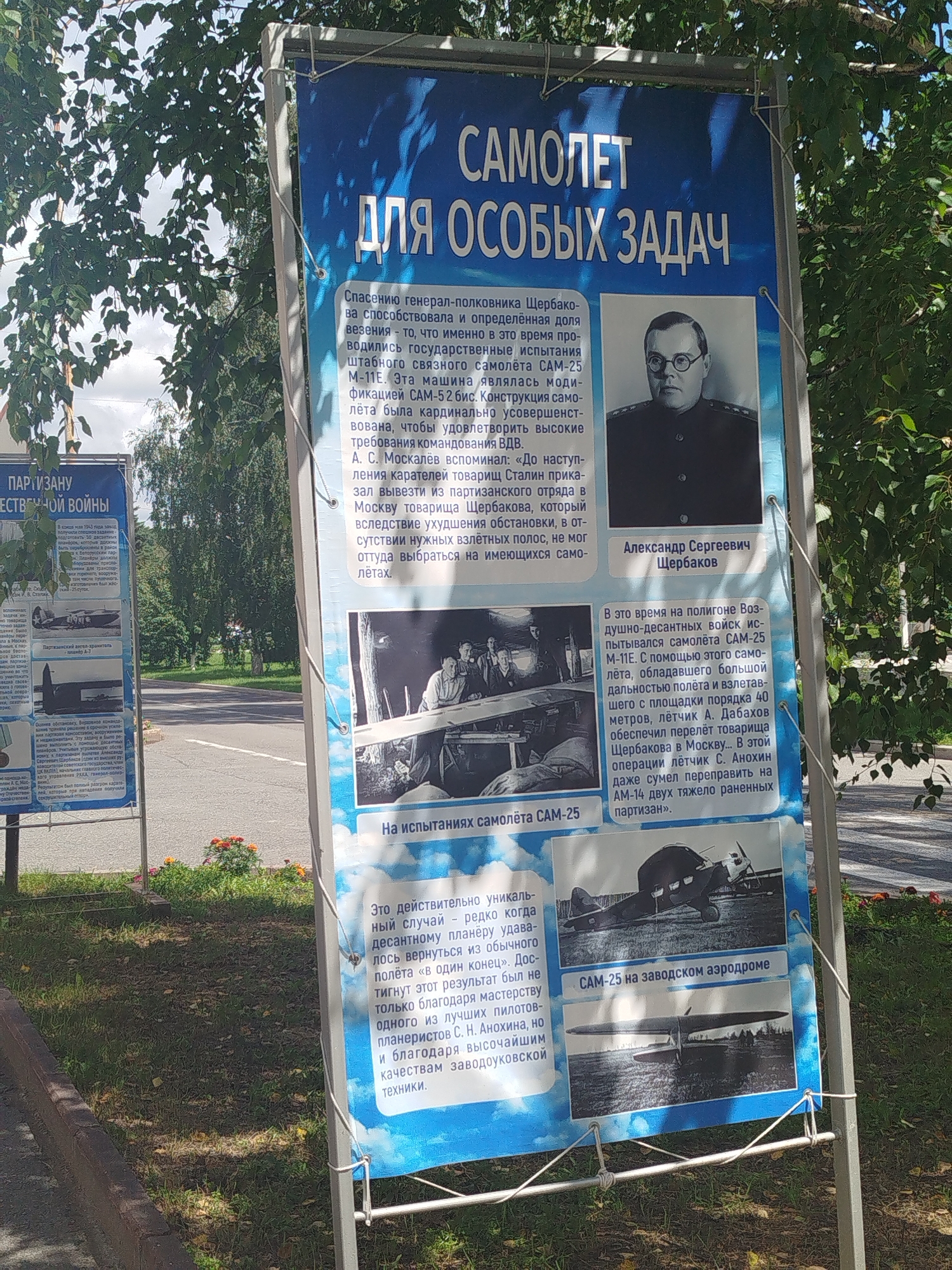 The image shows two large blue information boards with text and historical photographs, positioned outdoors in what appears to be a public space. The boards are likely part of an outdoor exhibition. In the foreground, the boards are mounted on metal poles, with some evidence of wear and tear on the edges. To the left of the main board, another partially visible board with similar content can be seen.
The main board, titled "САМОЛЕТ ДЛЯ ОСОБЫХ ЗАДАЧ" (Aircraft for Special Missions), features a portrait of a man in uniform, identified as Александр Сергеевич Щербаков (Alexander Sergeevich Shcherbakov). It also includes historical photographs of aircraft and people, accompanied by text in Russian. One photograph shows a biplane with the caption "Партизанский самолёт А-7" (Partisan aircraft A-7), and another shows two aircraft on an airfield with the caption "САМ-25 на заводском аэродроме" (SAM-25 at the factory airfield). The text describes the history and capabilities of these aircraft and the people involved in their development and operation, particularly during wartime.
The presence of the city name "Zavodoukovsk, Russia" is not directly visible in the image itself, but if this is a specific location, it implies the exhibition is situated within that city. The environment suggests an outdoor setting with trees and foliage, indicating a park or a public square. The lighting suggests it is daytime. The overall scene is informative, serving to educate viewers about a historical aspect of aviation.