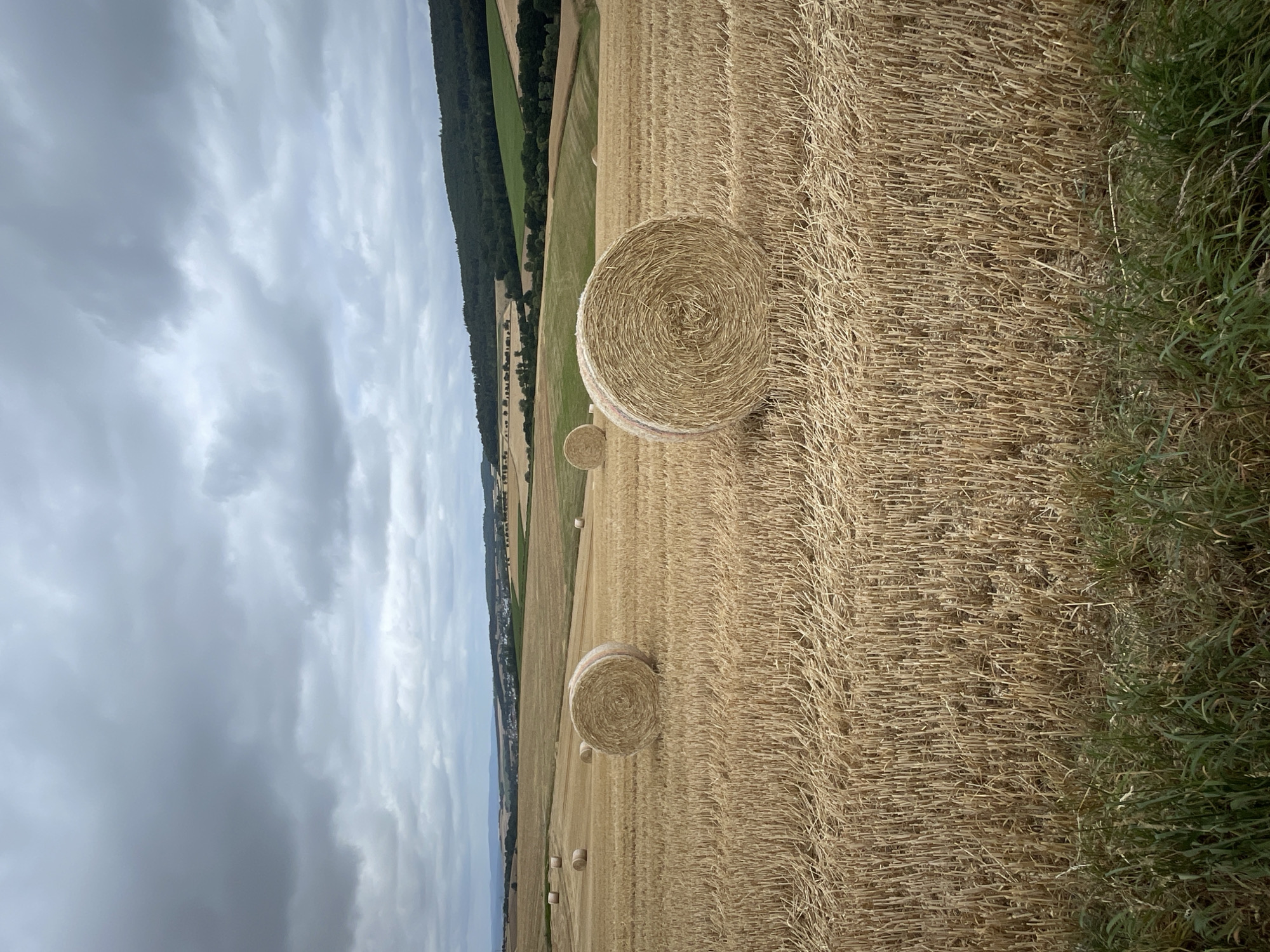 A landscape photograph captures a harvested wheat field under a cloudy sky. Several large, round bales of straw are scattered across the field. In the foreground, cut wheat stalks create a textured ground cover, with some green grass visible at the bottom edge. In the background, rolling hills are covered with trees and fields, and a small town is visible in the distance. The scene is likely in or around Asterode, Germany, given the architecture and landscape. There are no people or animals present.