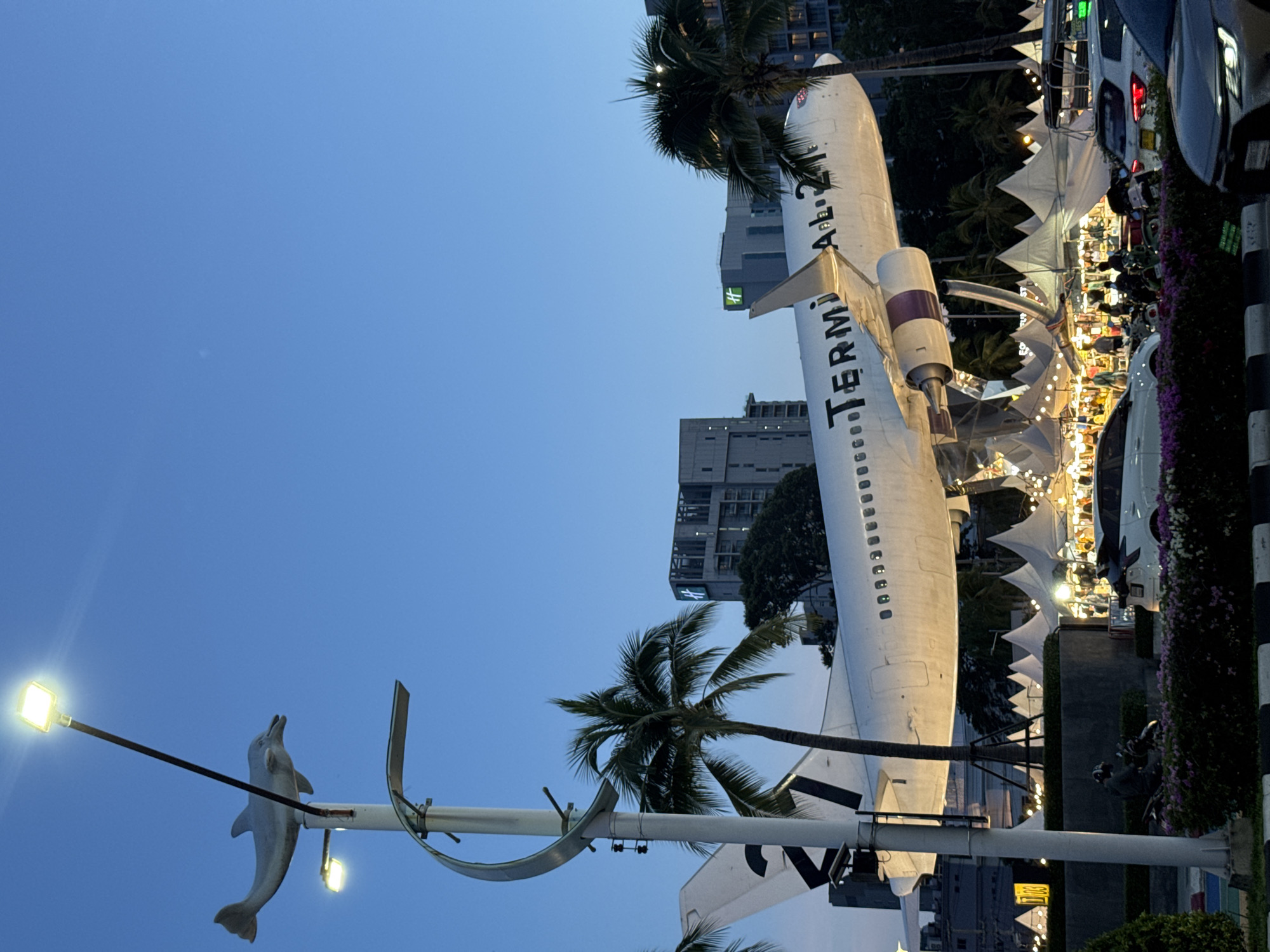 In Pattaya City, Thailand, a decommissioned airplane is displayed as an attraction, possibly at a market or event. The airplane, marked with "TERMINAL 21", is positioned horizontally, appearing to be supported by a structure, and is illuminated from below. In the foreground, a street light with a dolphin sculpture on top stands to the left of the frame. Palm trees surround the airplane, and behind it, several modern buildings are visible. A market with white tents and strings of lights is set up beneath the airplane. People are present, browsing the market, riding motorcycles, and there are vehicles on the road, including cars and a tuk-tuk. The sky is a clear, deep blue, indicating it is twilight.