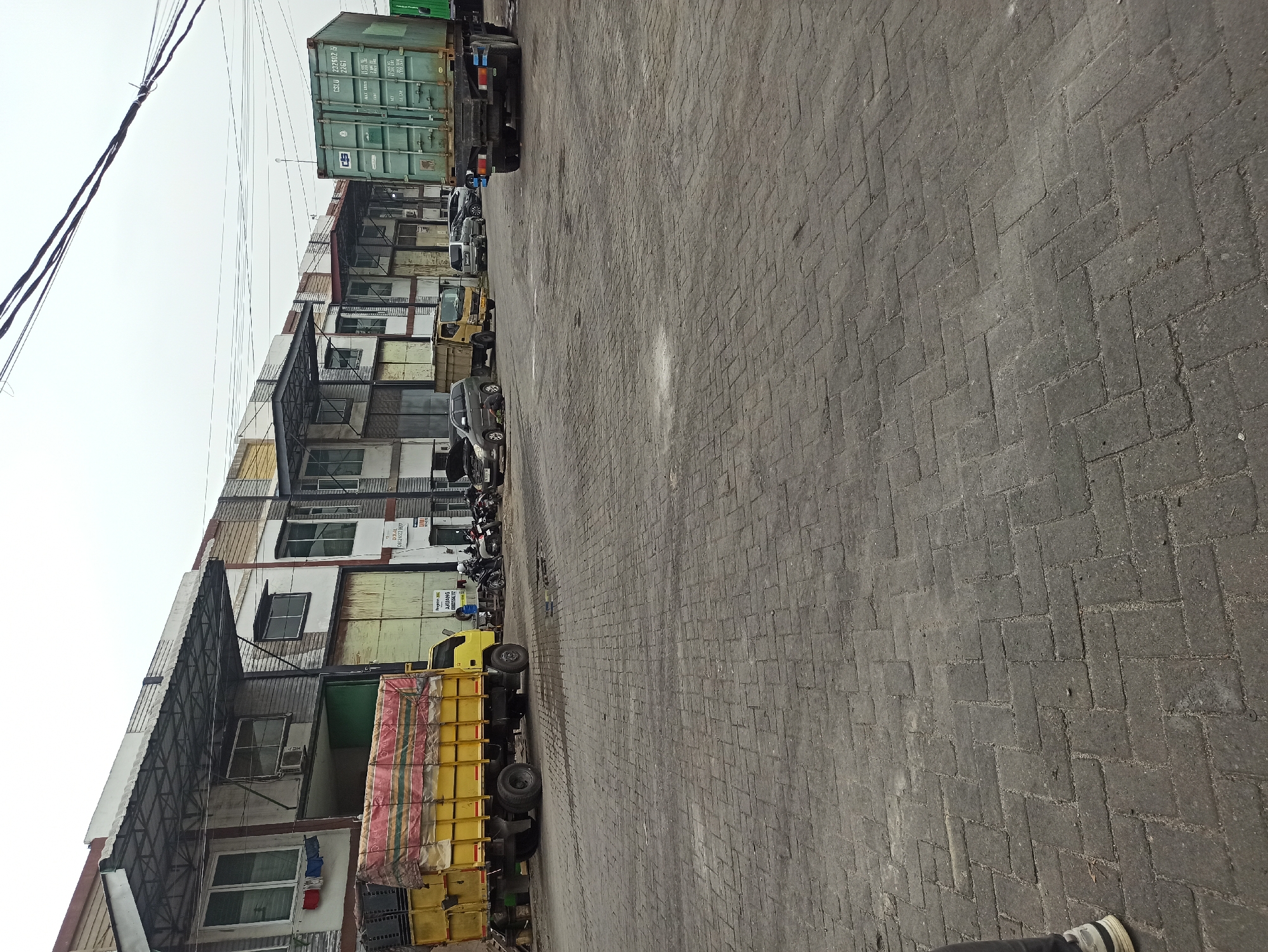 The image shows a row of commercial buildings in a city setting, likely an industrial or logistics area, under an overcast sky. The ground in the foreground is paved with interlocking grey bricks arranged in a herringbone pattern. To the left, a yellow truck with a covered cargo bed is parked. Further down the street, other vehicles are visible, including a green shipping container on a flatbed truck, a yellow truck, and several cars and motorcycles. Some of the buildings have exposed metal canopies over their entrances. A person's legs and feet, wearing white shoes and grey pants, are visible in the lower left corner of the image. The buildings appear to be multi-story structures with windows and visible air conditioning units. The overall impression is of a functional urban area with active commercial traffic.