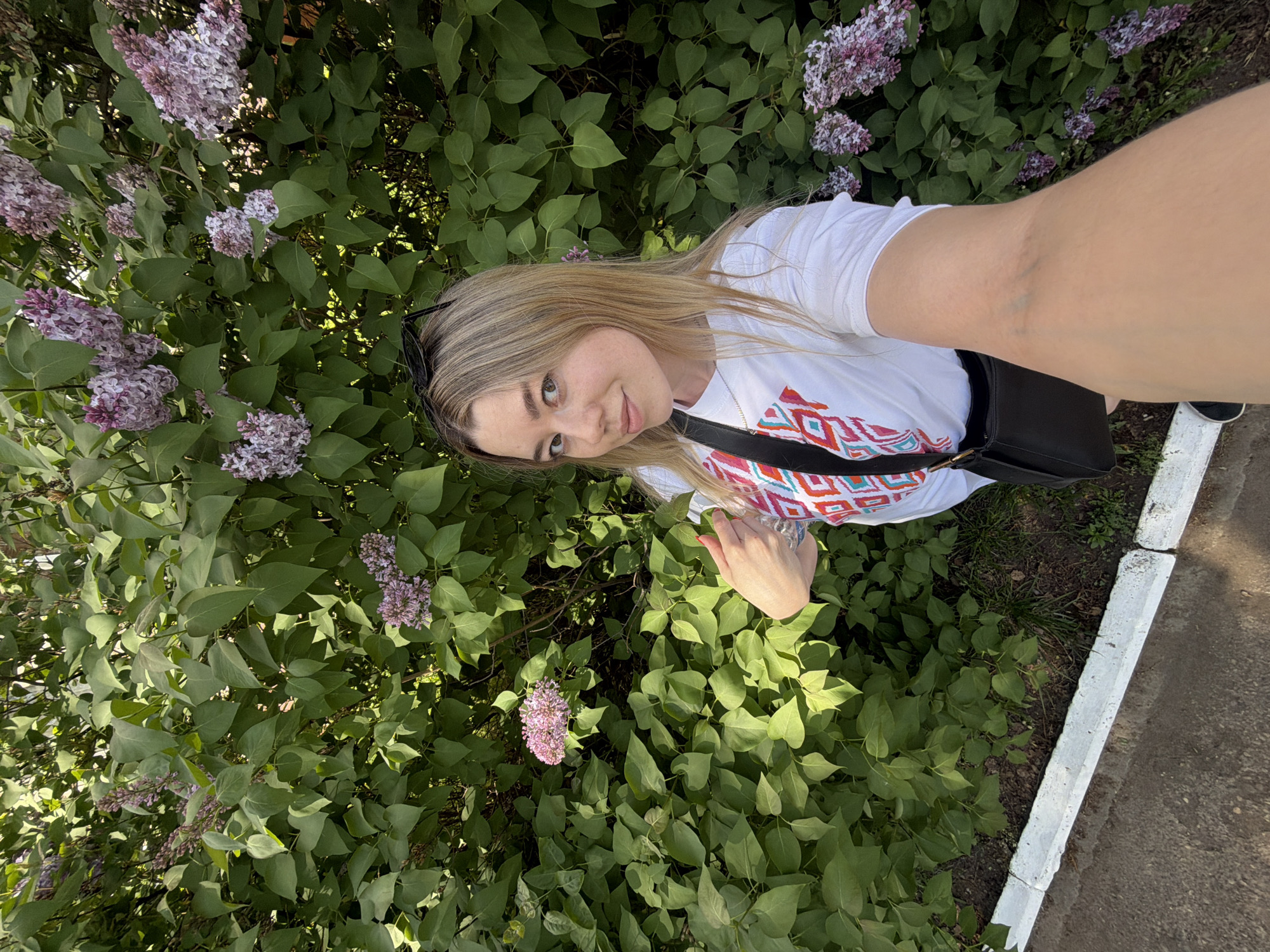 A woman stands next to a flowering lilac bush, holding a camera, likely taking a selfie. She is wearing a white t-shirt with a geometric pattern, a black shoulder bag, and sunglasses. Her hair is blonde. The lilac bush has purple and pink flowers. The location is likely in Chaikovsky, Russia, but specific details to confirm the exact location are absent. A concrete barrier is visible on the right side of the image.
