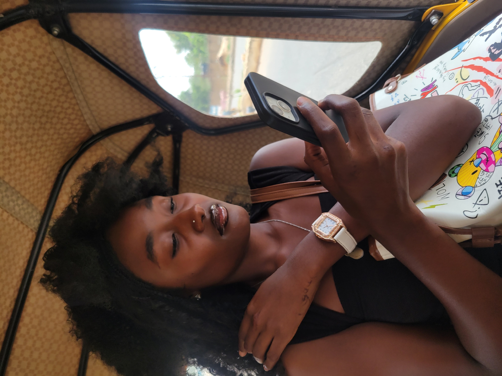The image shows a woman inside an auto rickshaw, likely in Jos, Nigeria, though this is not explicitly stated. She is dark-skinned with curly black hair, and wears a black top, a silver necklace, and a gold-colored watch with a white strap. She holds a black smartphone with multiple camera lenses in her hand and appears to be using it. Next to her is a white bag with cartoon graphics on it. The interior of the auto rickshaw has a checkered pattern and a visible window showing an out-of-focus street scene.