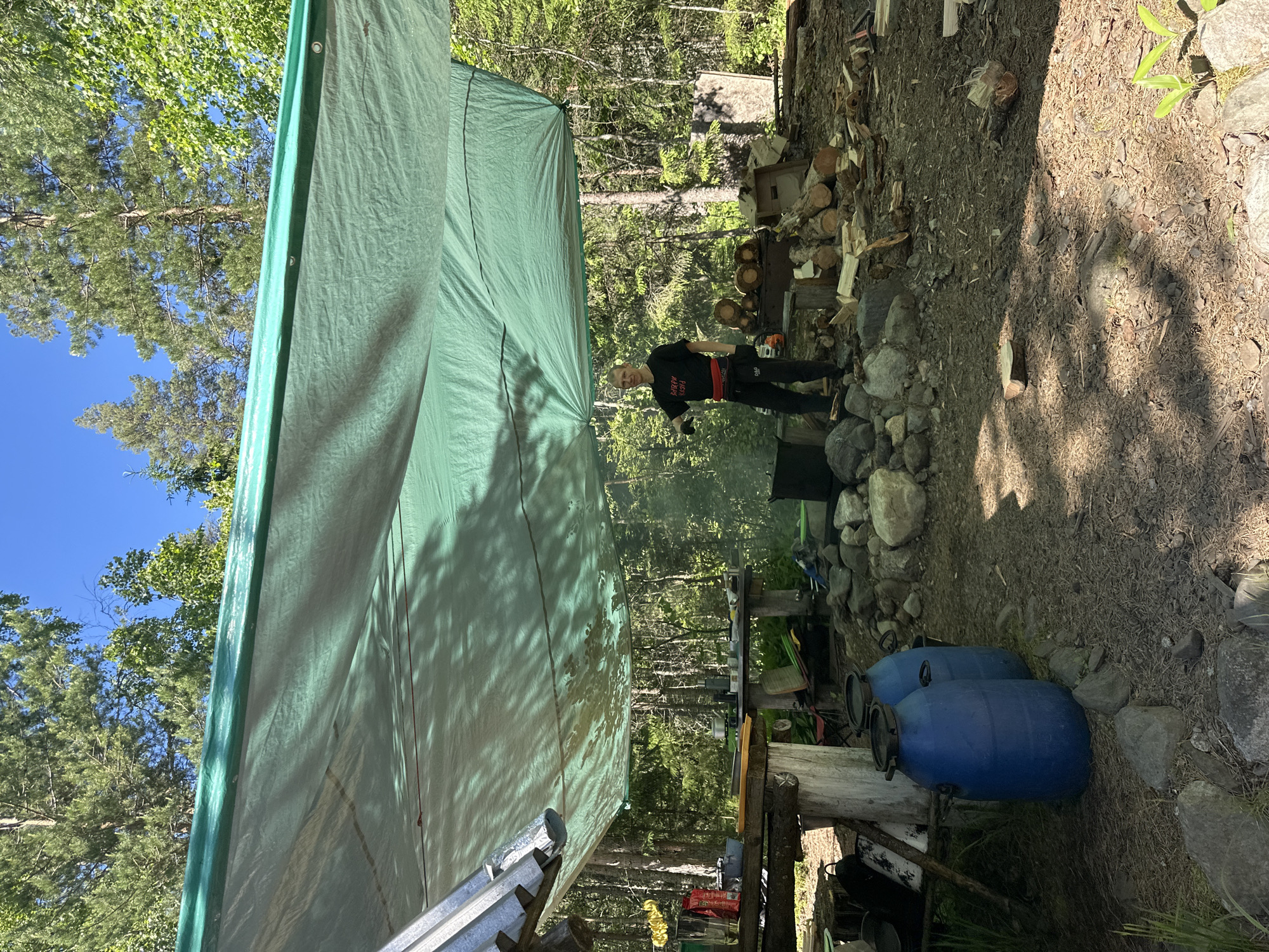 In Soanlakhti, Russia, a man in a black t-shirt and black pants stands under a large green tarp in a wooded area. He is holding a black object in his right hand and looking towards the camera. The tarp is supported by a wooden structure and appears to be providing shelter. In the foreground, there are two large blue barrels and various rocks scattered on the ground. Behind the man, there is a campfire with smoke rising, a pile of chopped firewood, and a wooden table with kitchen items. The environment is characterized by dense trees and a clear blue sky.