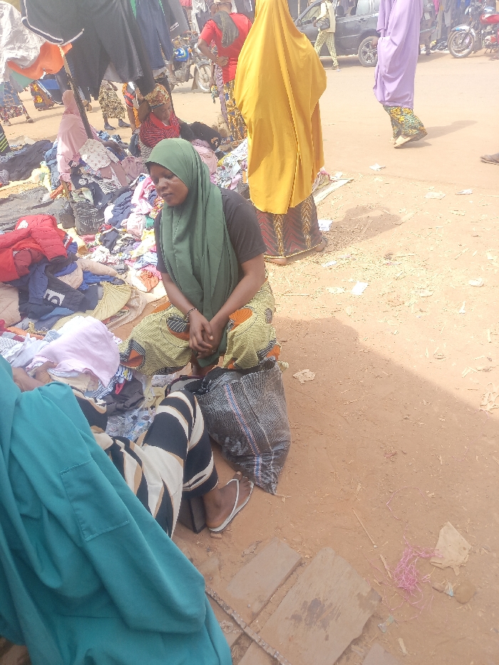 A woman wearing a green hijab and patterned pants sits on the ground in a bustling outdoor market. She has a black shirt and her hands are clasped in her lap. Nearby, a pile of clothes surrounds her. Other people wearing traditional clothing are milling around. A large bag rests near her. The ground is reddish-brown and slightly uneven. Garments hang above, creating a makeshift canopy.