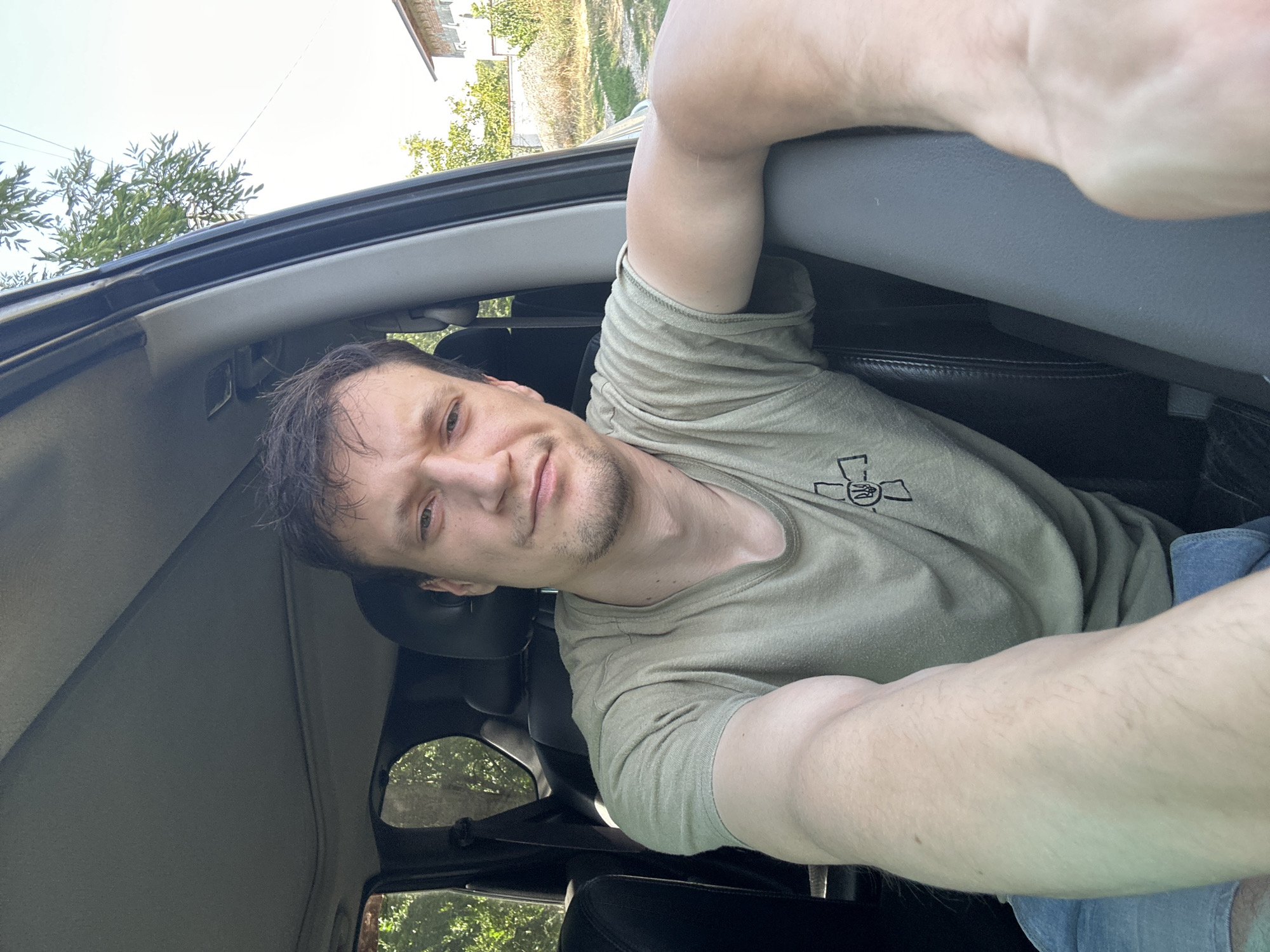A man with short brown hair and a light beard is sitting in the driver's seat of a car, taking a selfie. He is wearing a light olive-green t-shirt with a black emblem on the left chest and denim shorts. His left arm is extended towards the camera, resting on the car's interior. The background shows trees and a building, suggesting an outdoor setting. The car's interior is visible, including a black leather seat and a grey dashboard. The image appears to have been taken during the day, with natural sunlight illuminating the scene. The location is identified as Makiv, Ukraine.