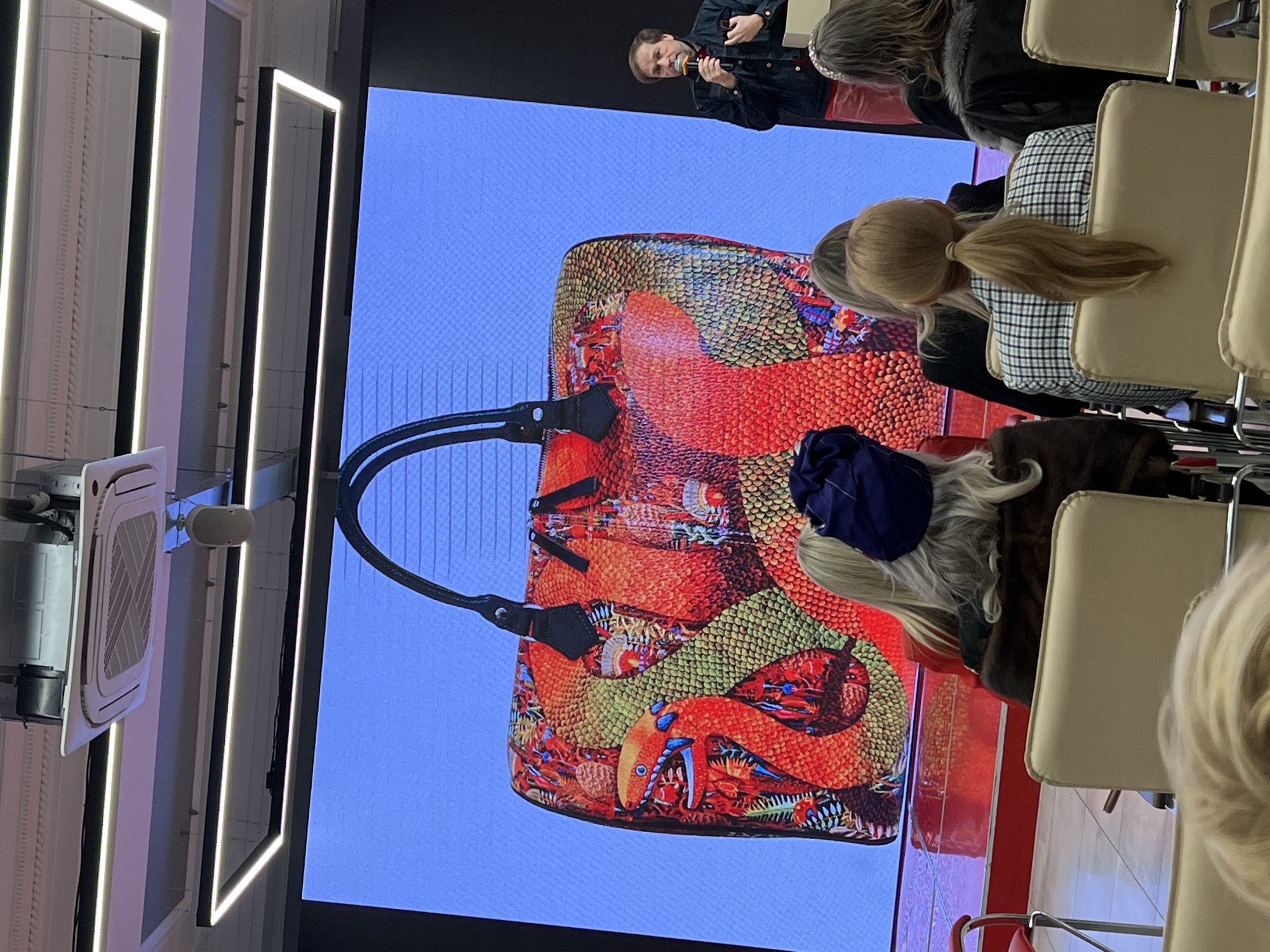 Captured indoors, seemingly in Saint Petersburg, Russia, the image presents a speaker addressing an audience in front of a screen showcasing a large handbag. The handbag features a unique, vibrant design dominated by orange and green tones, resembling scales and intricate patterns. The speaker, partially visible at the top, is holding a microphone, suggesting a presentation or talk. The foreground is filled with the backs of audience members seated on beige chairs. A ceiling-mounted air conditioning unit and minimalist light fixtures are noticeable in the background, adding to the modern interior of the location. The overall atmosphere suggests a formal event, possibly a fashion presentation or product launch.