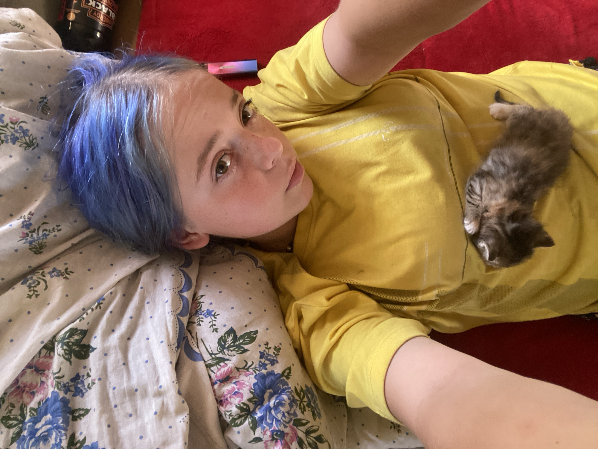 A young person with blue-dyed hair lies on their back, looking up at the camera. They are wearing a yellow t-shirt and are nestled against floral patterned bedding. A tiny kitten is curled up on the person's chest. The person's left arm is resting on the bedding, and their right arm is extended as if taking the selfie. The background is a red surface, possibly a bedspread or blanket. A black energy drink can is visible in the upper right corner. The scene appears to be indoors, in a bedroom. The location in Nova Praha, Ukraine is not discernible from the image.