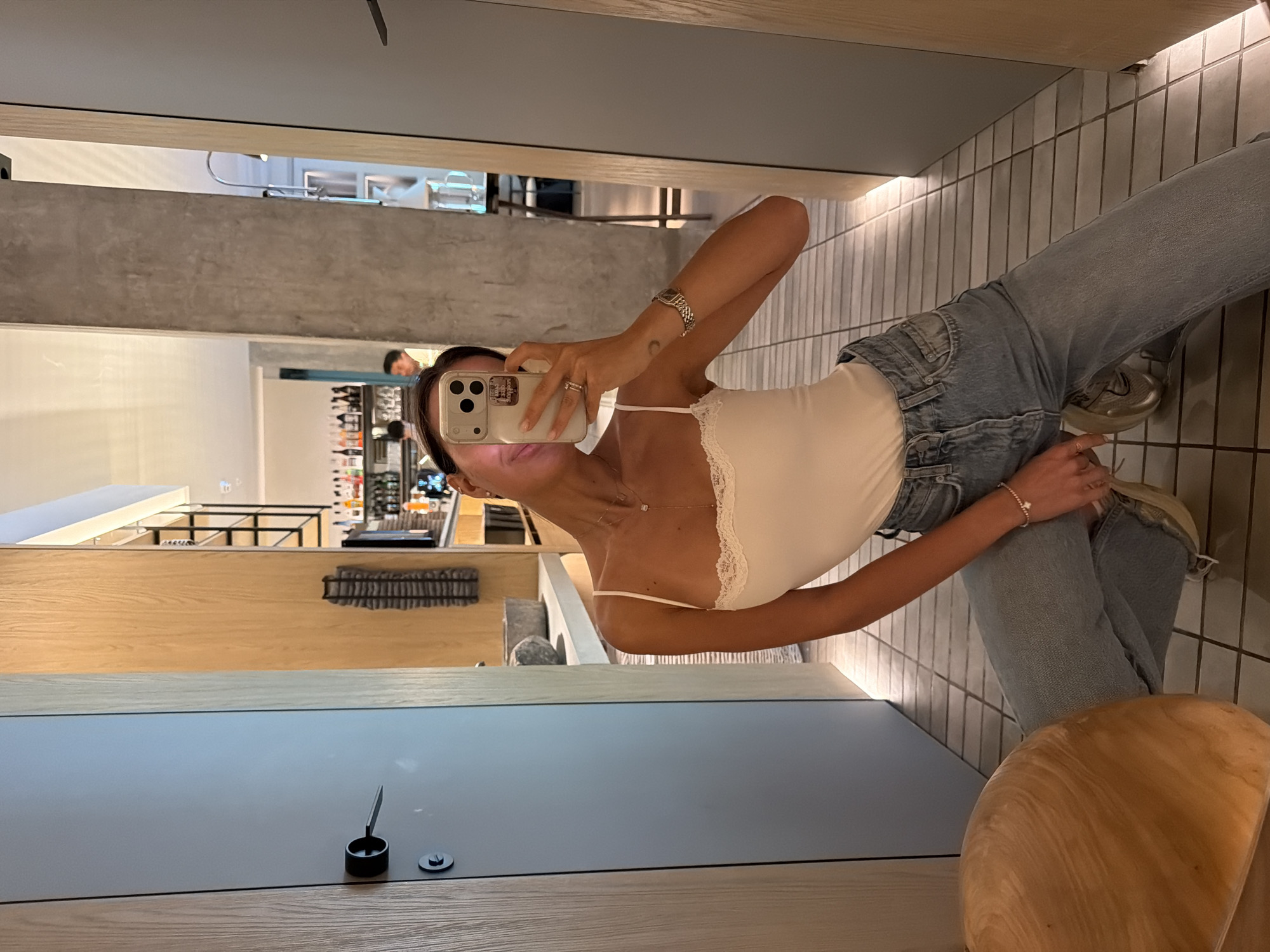 A woman stands in a modern commercial space, likely a restaurant or retail store, and takes a selfie using a smartphone. She is wearing a white top, light wash jeans, and light-colored sneakers. The room features a tiled floor and is furnished with wooden elements, including a table and a chair partially visible in the lower right corner. There is a view into another part of the establishment, showing shelves of products. A person is faintly visible in the background. The location is Krasnoyarsk, Russia.