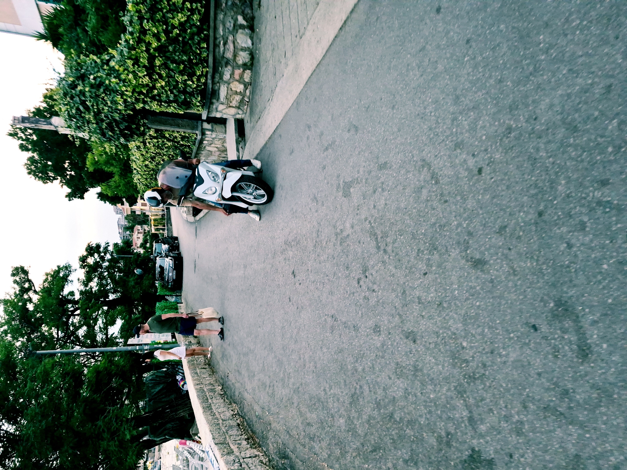 This media file, filmed in Đenovići, Montenegro, depicts a street scene with a focus on people and a scooter. The image is oriented vertically.
In the foreground, a white scooter is prominently featured. A person, wearing a helmet and a light-colored top with dark pants, is seated on the scooter, appearing to be either about to depart or having just arrived. Their posture suggests casualness, and the helmet indicates a concern for safety while riding.
Further in the background, two individuals are standing near a low stone wall adorned with lush greenery. One person, wearing a dark green t-shirt and shorts, stands upright, holding a bag. The other person, in a white top and shorts, is also standing, looking towards the street. Their casual attire and relaxed stances suggest a leisurely moment.
The setting appears to be a street in a residential or coastal area, with buildings visible in the distance. Trees with dense foliage line the left side of the road, creating a natural canopy. The road surface is asphalt, with a clear sidewalk or pedestrian path demarcated by a lighter colored material and a stone border.
The lighting suggests it is daytime, possibly late afternoon or early evening, given the soft illumination and the slightly muted colors. There are no visible signs of distress or urgency; the overall atmosphere seems calm and unhurried. The scene captures a slice of everyday life in a scenic location.