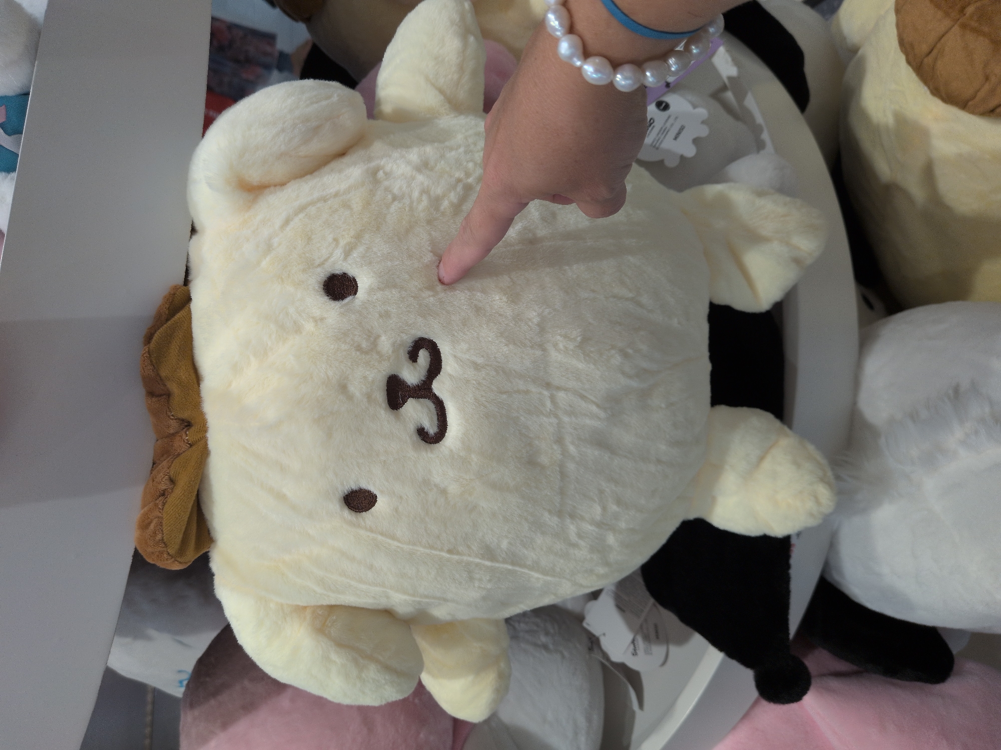 A large, plush, cream-colored stuffed dog, resembling the character Pompompurin, is the central subject of this image. The dog has two dark brown oval eyes, a small brown embroidered mouth, and is wearing a brown beret on its head. Its body is round and soft, with two rounded ears sticking out. A person's right hand, adorned with a pearl bracelet and a blue string, is extended towards the stuffed dog, with its index finger lightly touching the dog's cheek. The stuffed dog is situated on a white shelf, surrounded by other stuffed animals in varying colors, including pink, black, and white. The background suggests an indoor retail environment, possibly a toy store or a display area, with the overall impression being one of a playful and soft assortment of merchandise. This scene is located in Telford, United Kingdom.