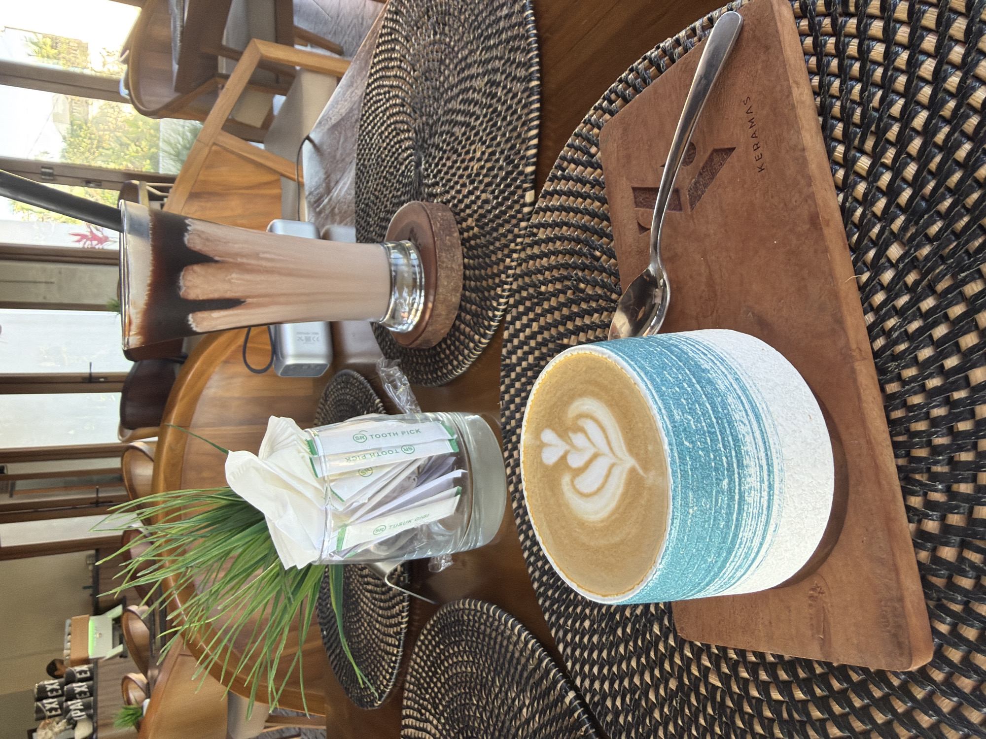 An overhead, close-up, eye-level shot shows a latte and a chocolate milkshake on a wooden table at a cafe in Blahbatuh, Indonesia. The latte is in a blue and white ceramic cup, decorated with latte art, and rests on a wooden coaster with the logo "KERAMAS" etched into it. A spoon is placed next to the cup. In the background, a tall glass of chocolate milkshake with a straw stands on a wooden base. A glass jar filled with individually wrapped toothpicks and some artificial greenery is situated behind the latte. The setting appears to be an outdoor or semi-outdoor restaurant with wooden chairs and tables visible in the background, suggesting a relaxed ambiance. The lighting is bright, indicative of daytime. No people are clearly visible in the frame, but the presence of food and drinks implies activity within the cafe.
