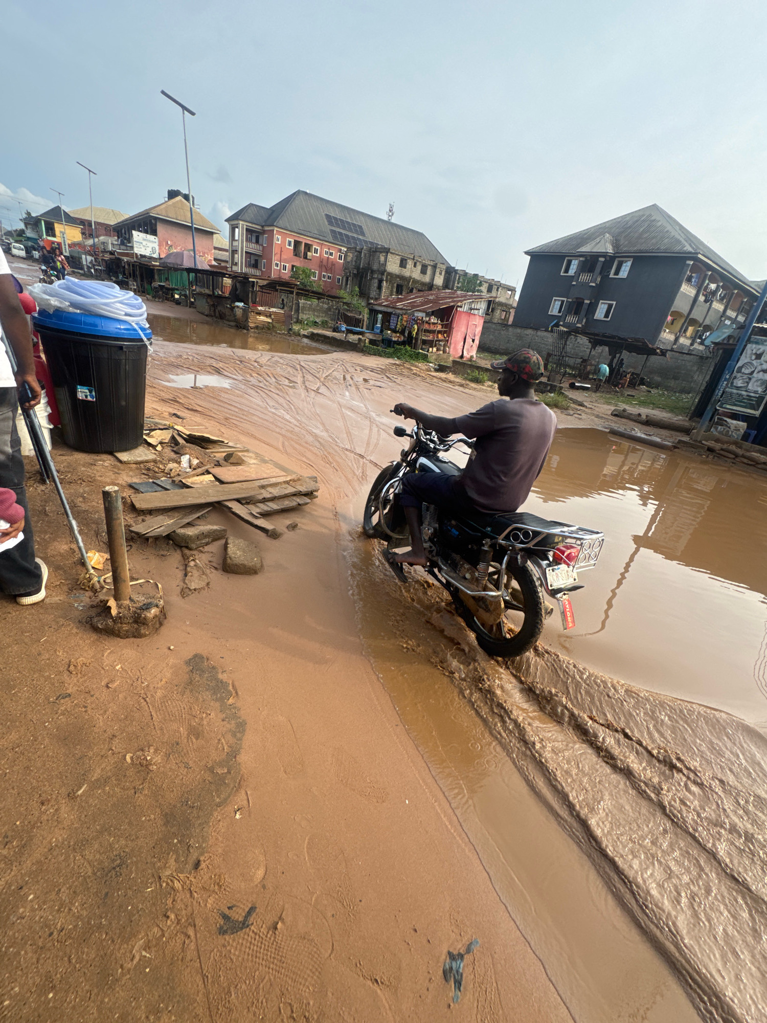 In Ihiagwa, Nigeria, a person rides a motorcycle through a flooded street. The water, mixed with mud, covers the road and appears to be flowing. Buildings line the street, some of which show signs of wear and tear. A large blue and black trash bin is visible on the left side of the image, and debris, including pieces of wood and rocks, is scattered along the edge of the road. The sky is overcast.