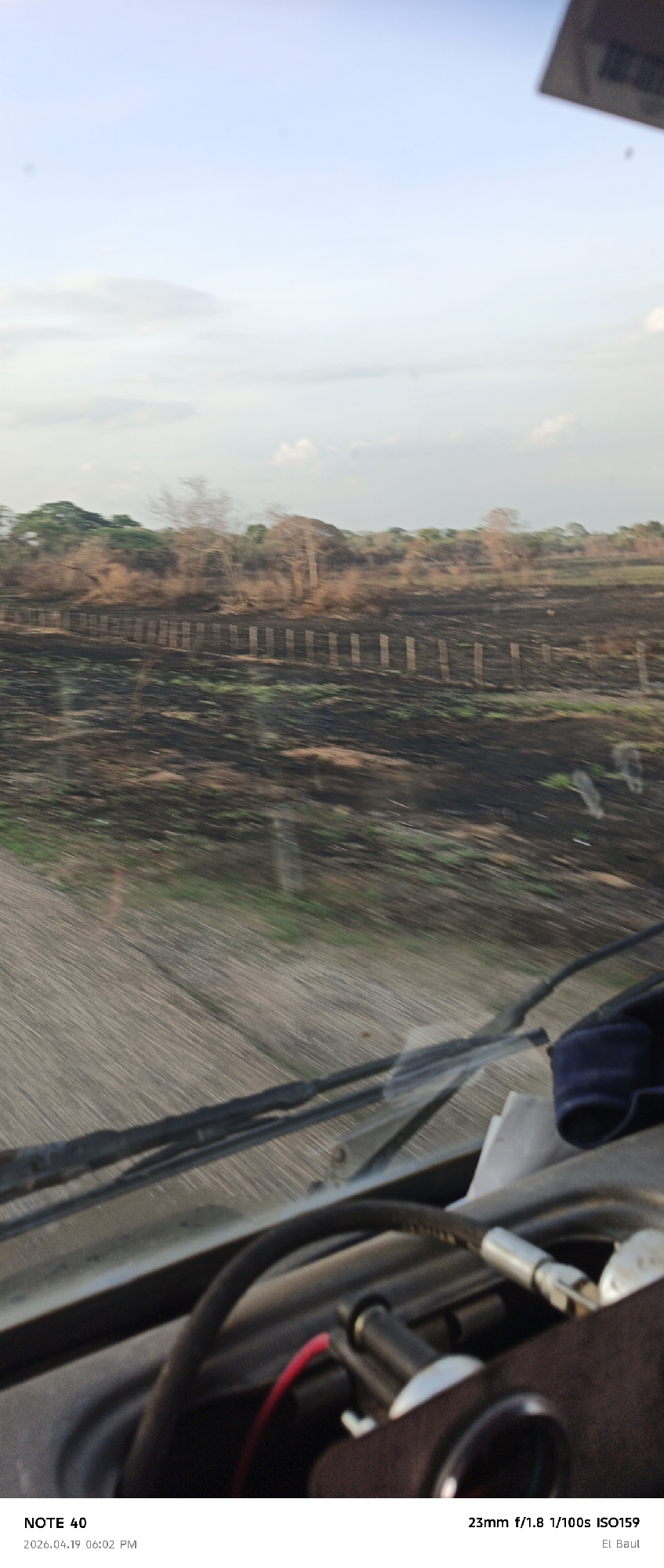 The image, taken from inside a vehicle, shows a landscape that appears to have been affected by a fire. The ground is mostly blackened, with some green vegetation beginning to emerge. A wooden fence line runs across the middle ground, separating the scorched earth from a line of trees in the background. The sky is overcast with light clouds. The foreground contains the dashboard of the vehicle, including windshield wipers and various wires and components. According to the image text, the photograph was taken in El Baul, likely located near La Cruz, Venezuela. The camera settings indicate a 23mm lens, f/1.8 aperture, 1/100s shutter speed, and ISO 159. The image capture date is noted as April 19, 2026.