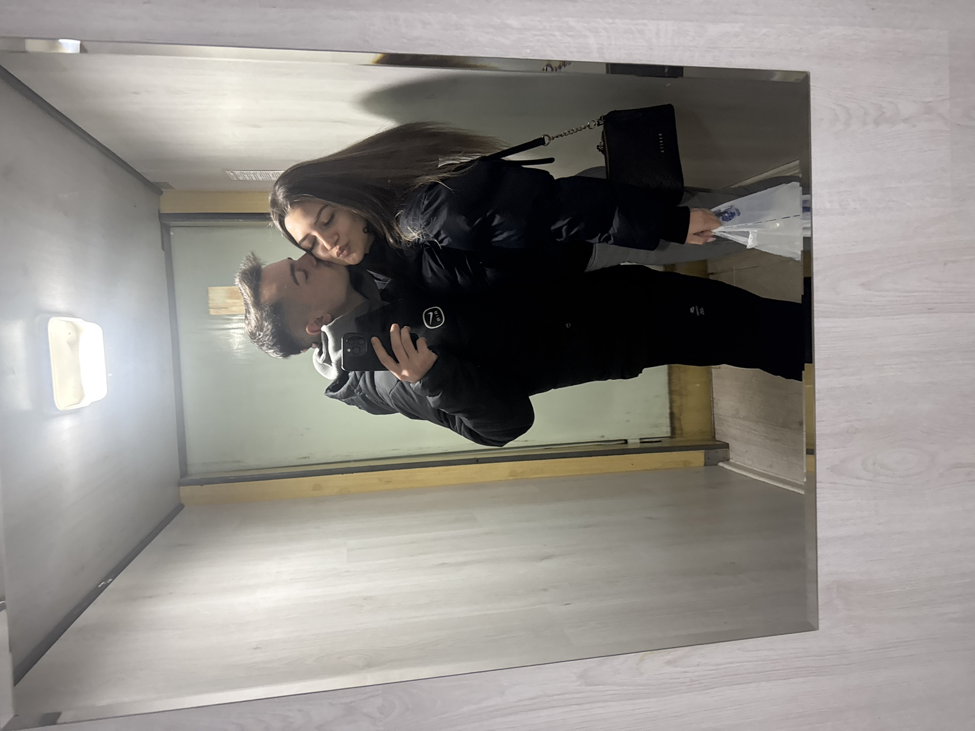 A young couple are reflected in a mirror. The man is holding a phone and taking a selfie as he kisses the woman's cheek. The woman is holding a shopping bag and a purse. They are in a narrow corridor with grey walls, possibly in an apartment building or shop. The location is unknown, so I can not determine if it is in Sofia, Bulgaria.