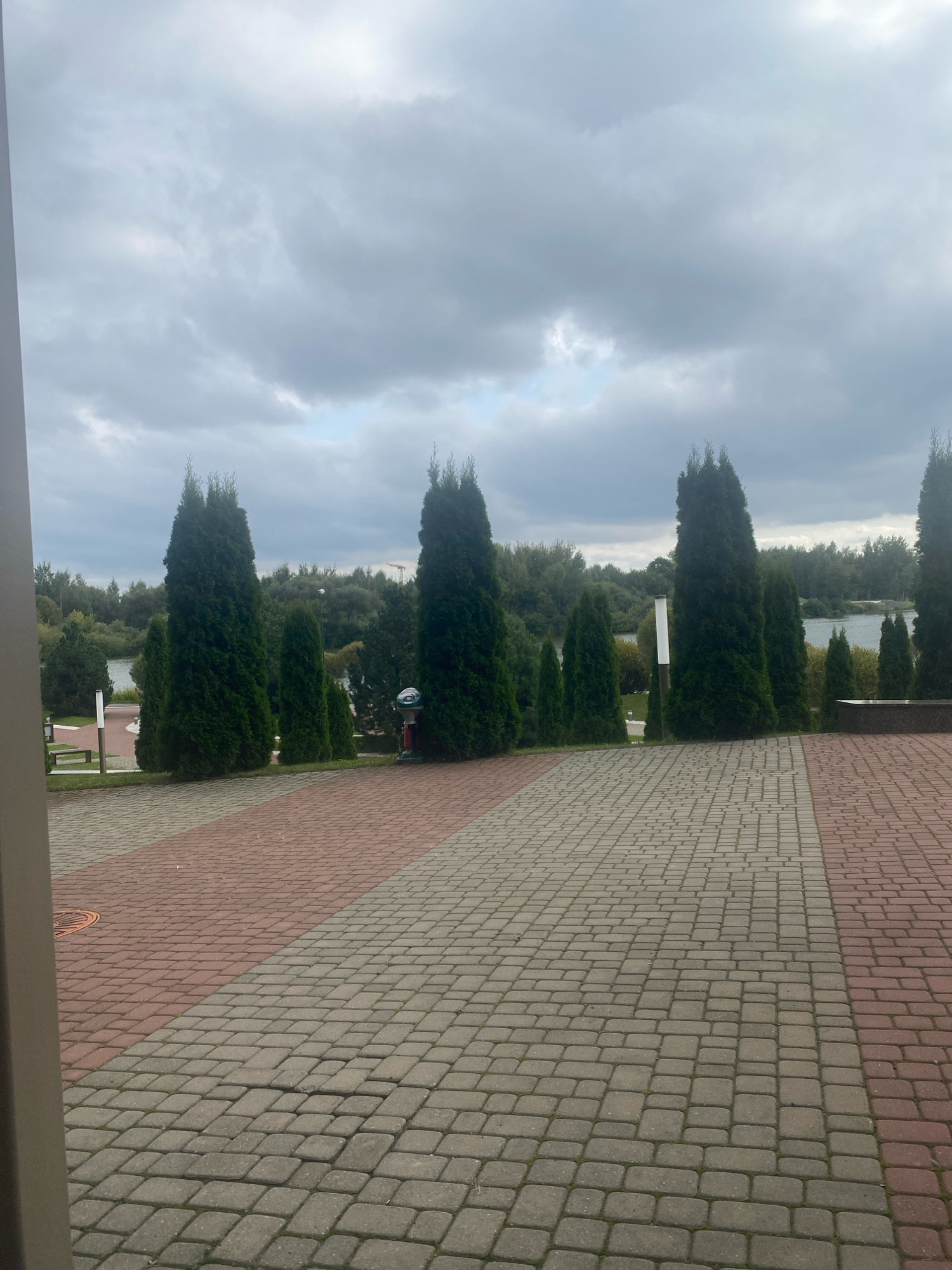 The image shows a paved walkway with a pattern of red and gray interlocking pavers. The walkway leads to a body of water lined with trees, and several tall, dark green evergreen trees are planted in a row along the edge of the walkway. The sky is overcast with gray clouds. In the distance, a faint image of a crane is visible, suggesting construction. There are no people or animals visible in the image. The scene appears to be a public park or recreational area.