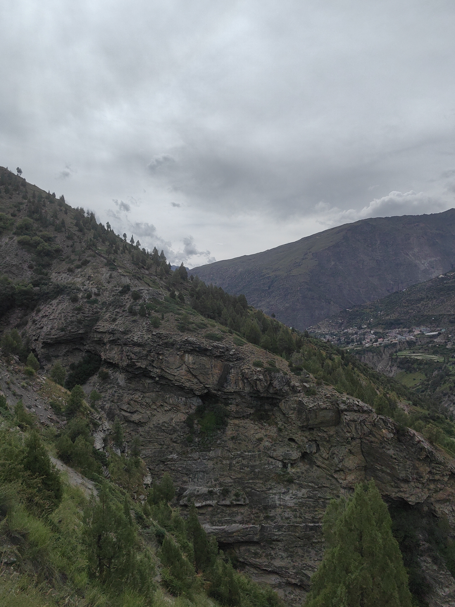 This media file captures a scenic view of a mountainous landscape in Keylong, India. The image focuses on a steep, rocky mountainside dotted with sparse vegetation, including numerous coniferous trees. In the background, a larger, more distant mountain range is visible, with a small village nestled in the valley between the peaks. The sky is overcast with heavy, gray clouds, suggesting a cloudy or possibly rainy day. The overall mood is somewhat somber, reflecting the dramatic and rugged terrain under the muted sky. There are no visible people, objects, or text in the image. The time of day appears to be daytime, but the cloudy conditions obscure any direct sunlight. The location cues, such as the village and the characteristic mountainous terrain, are consistent with the region of Keylong.