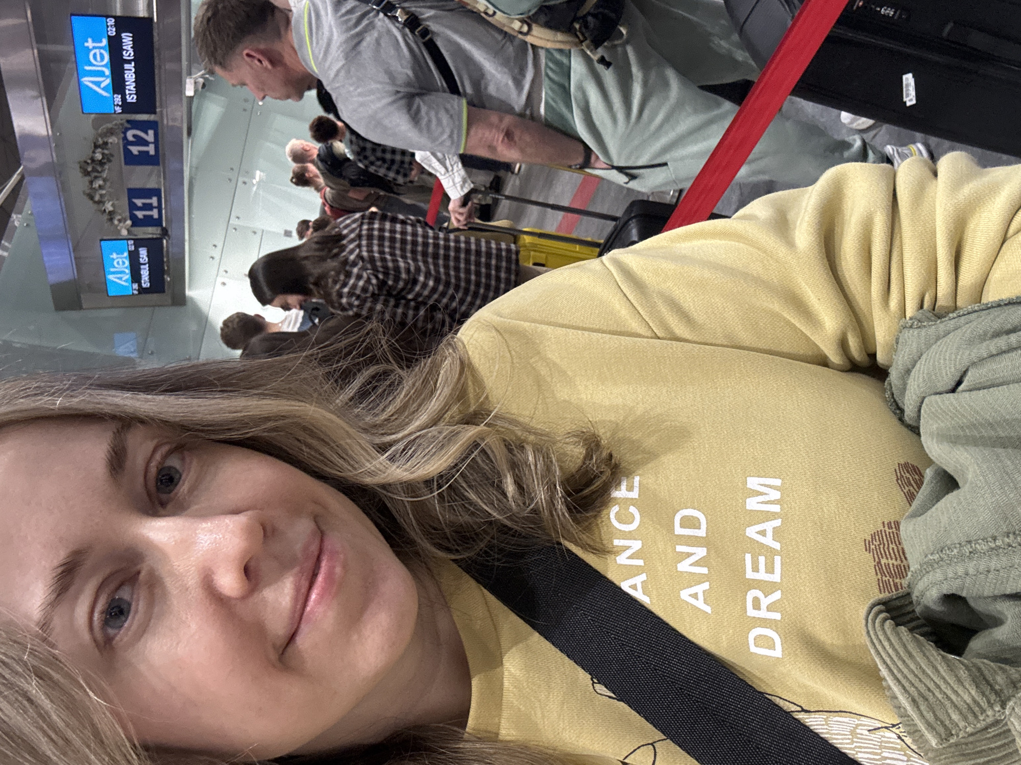The image captures an indoor airport gate scene, featuring a woman in the foreground taking a selfie, with a queue of passengers and flight information displays in the background.
**Location:** The digital display boards clearly indicate "AJet" flights to "ISTANBUL (SAW)" (Sabiha Gökçen International Airport, Istanbul, Turkey) with flight number "VF 232" and a scheduled time of "02:10" at gates "11" and "12". There is no visual information within the image to suggest the location is in the city of Saint Petersburg, Russia. The flight details indicate an airport connecting to or from Istanbul, Turkey.
**People:**
* **Foreground:** A fair-skinned woman with light-colored eyes and wavy, light brown hair is positioned in the lower-left, taking a selfie. She has a faint smile and looks directly at the camera. She wears a mustard-yellow long-sleeved sweatshirt, with the white text "ANCE AND DREAM" (likely part of "DANCE AND DREAM") visible on her chest. A black bag strap crosses diagonally over her torso. Part of a light green, ribbed garment is visible in the lower left, possibly held in her arm.
* **Background:** A queue of approximately 8-10 individuals, predominantly adults, extends from the middle-ground into the distance. Most individuals are facing away from the camera or in profile.
* A woman in a brown and black plaid shirt stands in the middle-ground, holding the handle of a yellow roller suitcase.
* On the far right, a man in a grey t-shirt with a neon green trim on the collar and light green sweatpants is visible from the back and side, carrying a dark backpack and appearing to look towards the right.
* Other individuals in the queue are casually dressed, some pulling black roller suitcases.
**Objects and Environment:**
* The setting is a modern airport terminal with light-colored walls, possibly composed of glass or polished panels.
* Two blue digital display screens are mounted above the queue, both displaying the "AJet" logo, flight number "VF 232", destination "ISTANBUL (SAW)", and time "02:10".
* Below these screens, two blue signs clearly mark "11" and "12" as gate numbers.
* A white floral decoration is affixed to the wall between gate numbers 11 and 12.
* Red retractable belt barriers delineate the queuing area on the grey floor.
* Various pieces of luggage, including black and yellow roller suitcases, are visible on the floor within the queue.