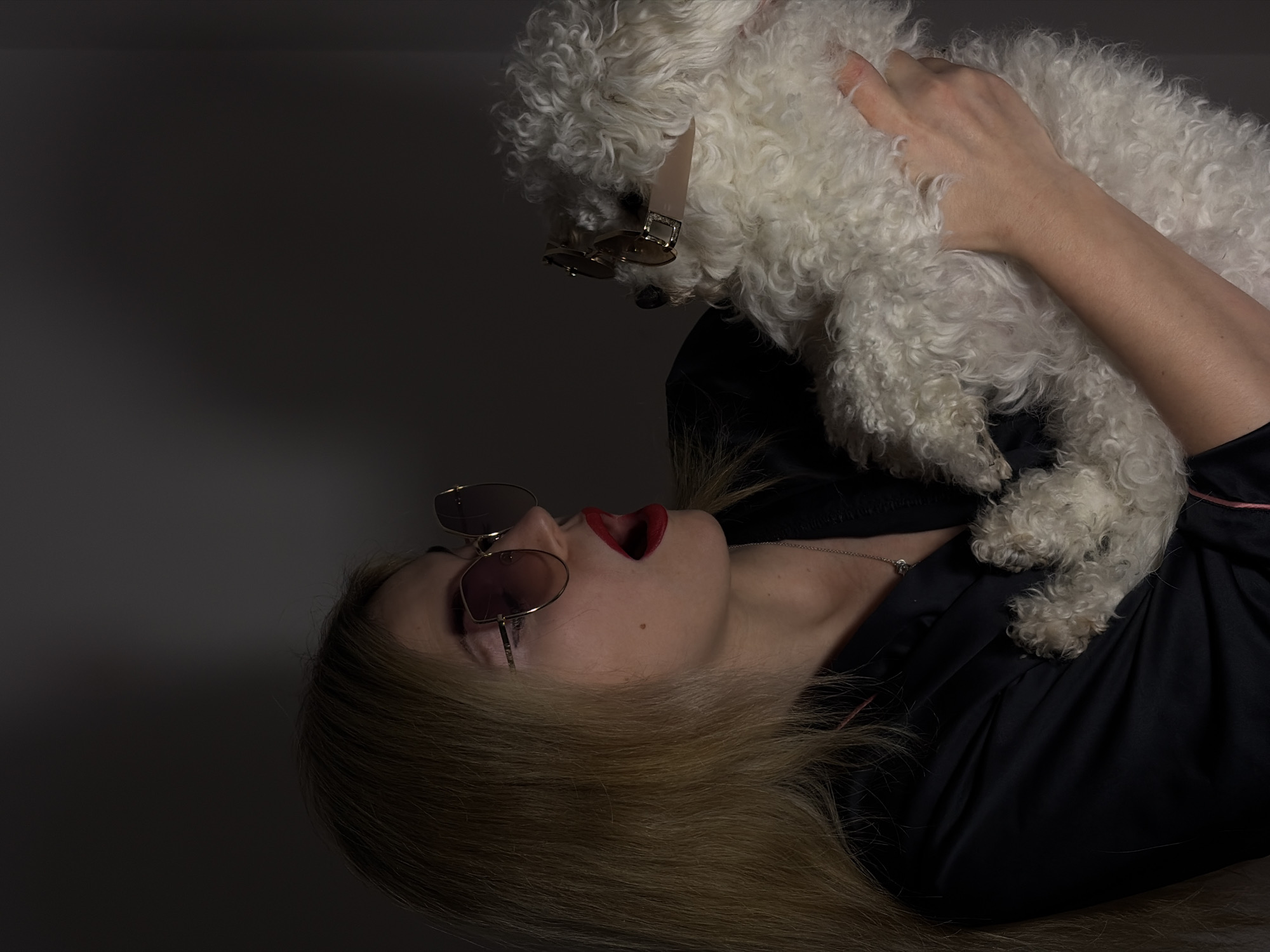 A person with blonde hair, wearing sunglasses and red lipstick, lies tilted to the right. The person looks upwards towards a fluffy white dog, also wearing sunglasses. The person is wearing a dark colored garment. The location is not discernible from the image.