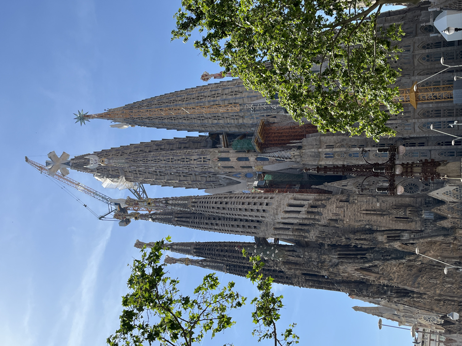 The image displays a partial view of the Sagrada Familia basilica in Barcelona, Spain, under a clear blue sky. The monumental, intricately designed structure dominates the frame, showcasing its characteristic towering spires and complex architectural details.
Several of the basilica's iconic towers are visible, rising majestically. One prominent tower features a multi-pointed star at its apex, while another adjacent tower is topped with a distinct cross-like finial. The stone work on the towers exhibits elaborate carvings, numerous arched windows, and sculptural elements, highlighting the unique Gothic-Art Nouveau style.
Evidence of ongoing construction is apparent, with at least two large cranes positioned near the towers, and some scaffolding or protective coverings visible on parts of the facade, underscoring the basilica's continuous development.
In the foreground, vibrant green foliage from trees partially frames the view, adding a natural contrast to the stone edifice. The bright sunlight suggests it is daytime, likely midday or early afternoon, with clear and sunny weather. The vivid blue sky provides a serene backdrop to the grand architectural masterpiece. No discernible text is visible on the building or in the surrounding environment.