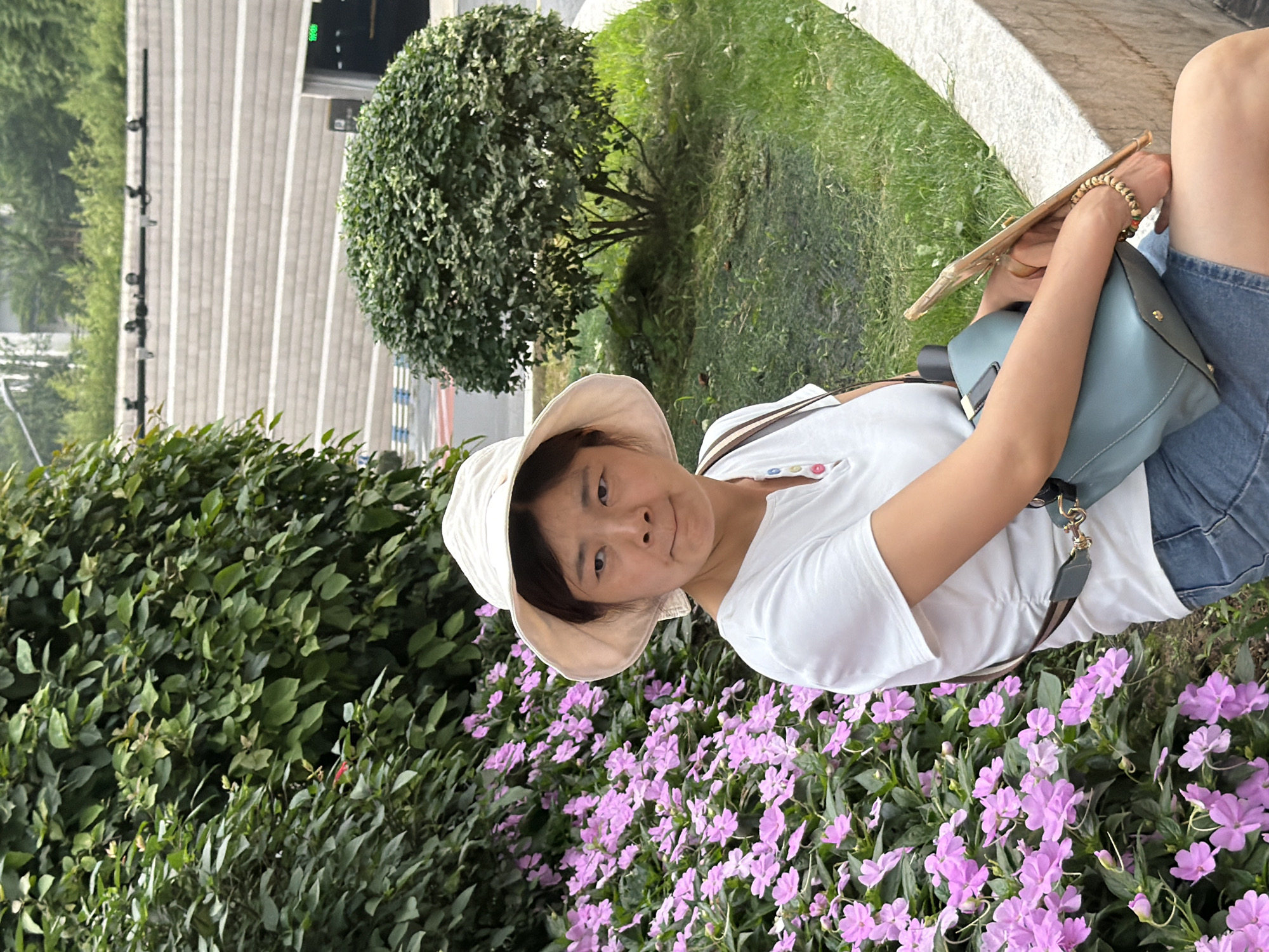 A woman wearing a white sun hat and a white polo shirt sits on a stone bench in front of a large cluster of pink flowers. She is wearing denim shorts and has a light blue purse slung over her shoulder. She is holding a wooden stick in her right hand and a beaded bracelet on her left wrist. In the background, there is a trimmed green bush and a terraced stone wall. The scene appears to be located in Yuzhong District, China.