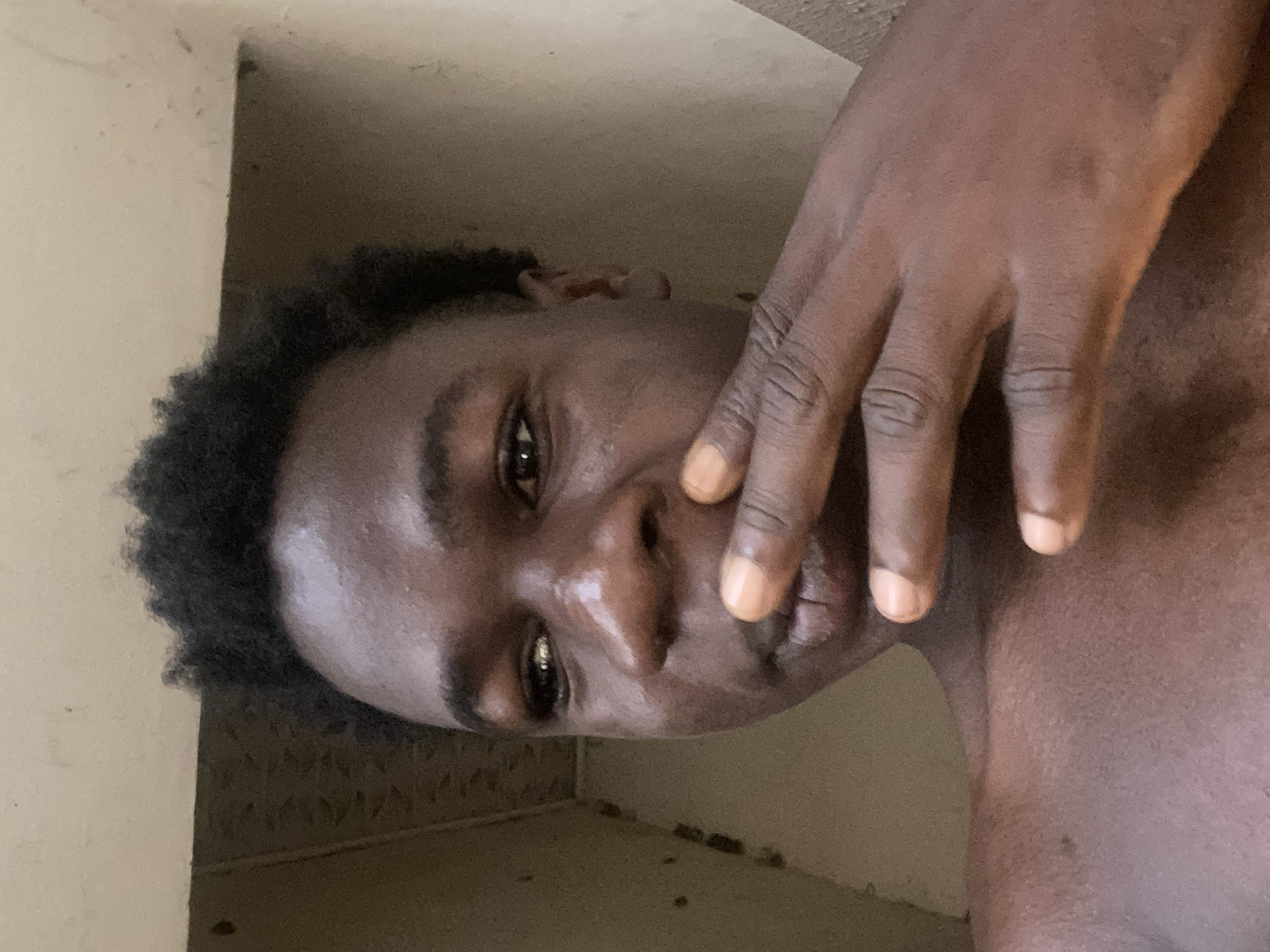The close-up, portrait-style image features a person of dark complexion, presumably male, with dark, curly hair. The individual's right hand is positioned over their face, partially obscuring the mouth and nose, with the visible eye looking directly towards the camera. The eye appears calm or neutral, contributing to a pensive or casual expression. The person's skin has a noticeable sheen, possibly due to natural oils or the specific lighting conditions. The fingernails on the visible hand are short, neatly trimmed, and painted a light, subtle peach or orange color.
The setting appears to be indoors, against a plain, light-colored wall that forms a corner in the background. The lighting is diffused and somewhat subdued, creating soft shadows. No distinct objects are visible in the background. The primary activity is posing for the photograph. There is no visible text within the image. The location context for this image is Mararaba, Nigeria.
