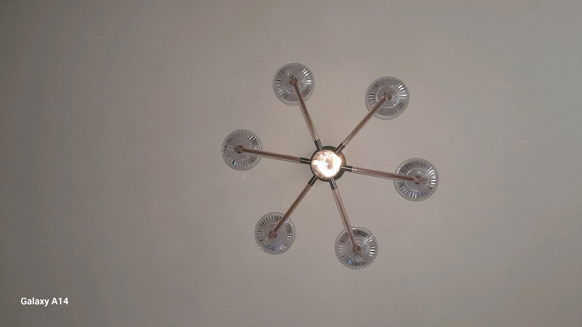 A low-angle shot captures a modern, star-shaped chandelier against a neutral ceiling. The chandelier features six arms extending from a central light fixture. Each arm is topped with a clear, circular glass shade with a ridged texture, catching the light and creating a subtle sparkle. The arms themselves appear to be made of a metallic material, possibly with a brushed finish, and are accented with dark connectors. The central light is on, emitting a warm glow. The "Galaxy A14" watermark is present in the bottom left corner. The scene is indoors, with the focus on the ceiling-mounted light fixture.