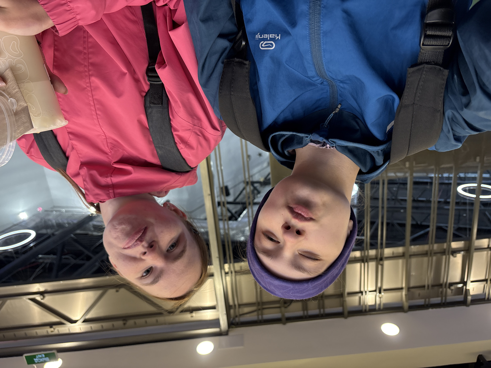 Two fair-skinned individuals are photographed from a low angle, giving the impression they are looking down at the viewer. The person on the left is wearing a pink jacket and a backpack. They are holding a drink in a clear plastic cup. The person on the right is wearing a blue jacket and a purple headband, also sporting a backpack. The background reveals an interior space with a metal ceiling structure, circular light fixtures, and an exit sign. The location appears to be a public space, possibly a transportation hub or commercial building in Perm, Russia.