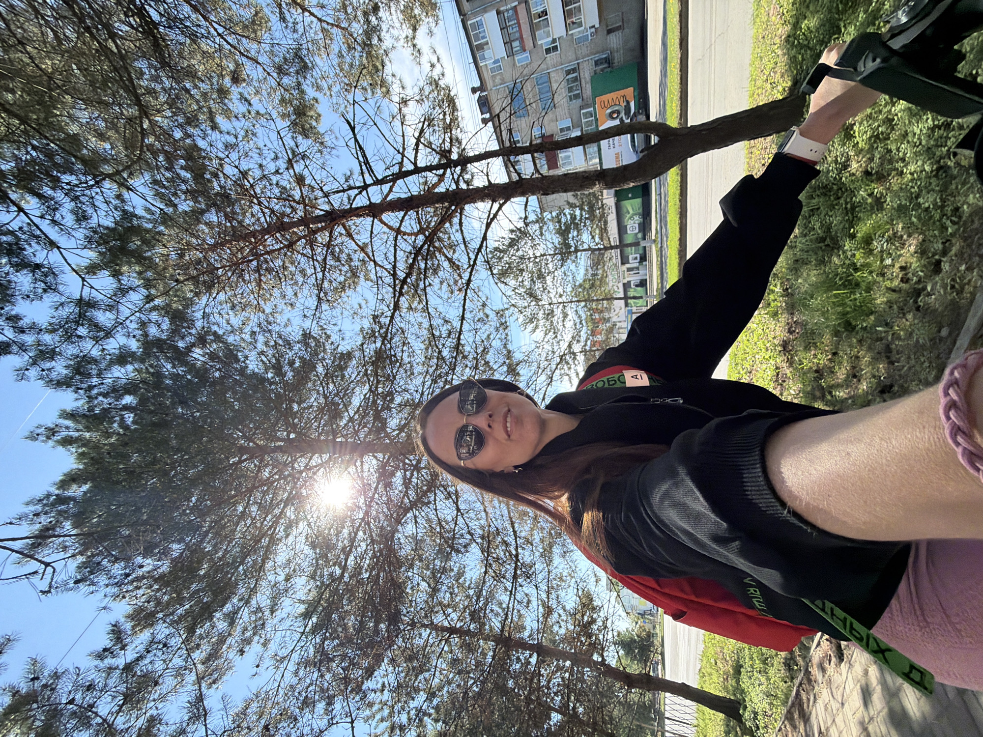 A medium shot shows a woman facing the camera while outdoors, likely in Birobidzhan, Russia, based on Cyrillic text on nearby buildings. The perspective suggests the photo was taken at an upward angle. The woman wears sunglasses, a black sweatshirt with red and green accents, and pink clothing. She is partly obscured by tree branches against a blue sky with visible sunlight. In the background, a multi-story residential building is seen, along with green foliage along the roadside. She is grasping the handles of a stroller or similar apparatus.