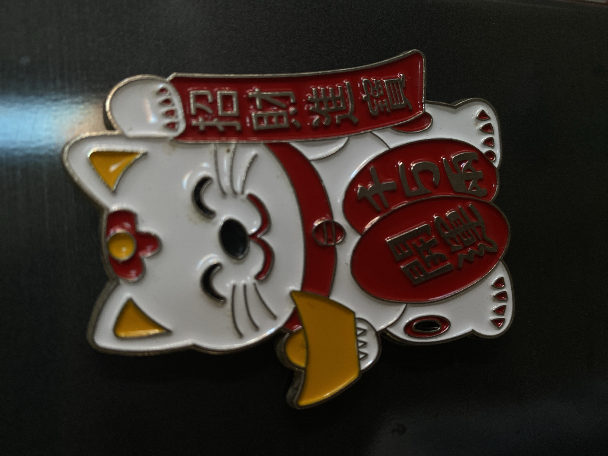 A white Maneki-neko, or lucky cat, charm is displayed against a dark, possibly black, background. The cat is depicted with its right paw raised, holding a red banner with Chinese characters that appear to read "招財進寶" (zhāo cái jìn bǎo), meaning "attract wealth and treasure." The cat wears a red collar with a gold circular ornament. In its left paw, it holds a golden sycee, a type of Chinese gold or silver ingot used as currency. Two large red oval shapes are placed on the cat's chest, each with Chinese characters. The left oval shows "開運" (kāi yùn), meaning "good luck" or "fortune." The right oval shows "招財" (zhāo cái), meaning "attract wealth." The cat has a smiling expression with closed eyes and whiskers. The charm is detailed with red and yellow accents on its ears and a red flower on its head. The background is dark and somewhat reflective, showing a hint of blue light at the top. The image is taken from a top-down perspective, with the charm lying flat. There is no indication of the city of North Kuta, Indonesia within the image.
