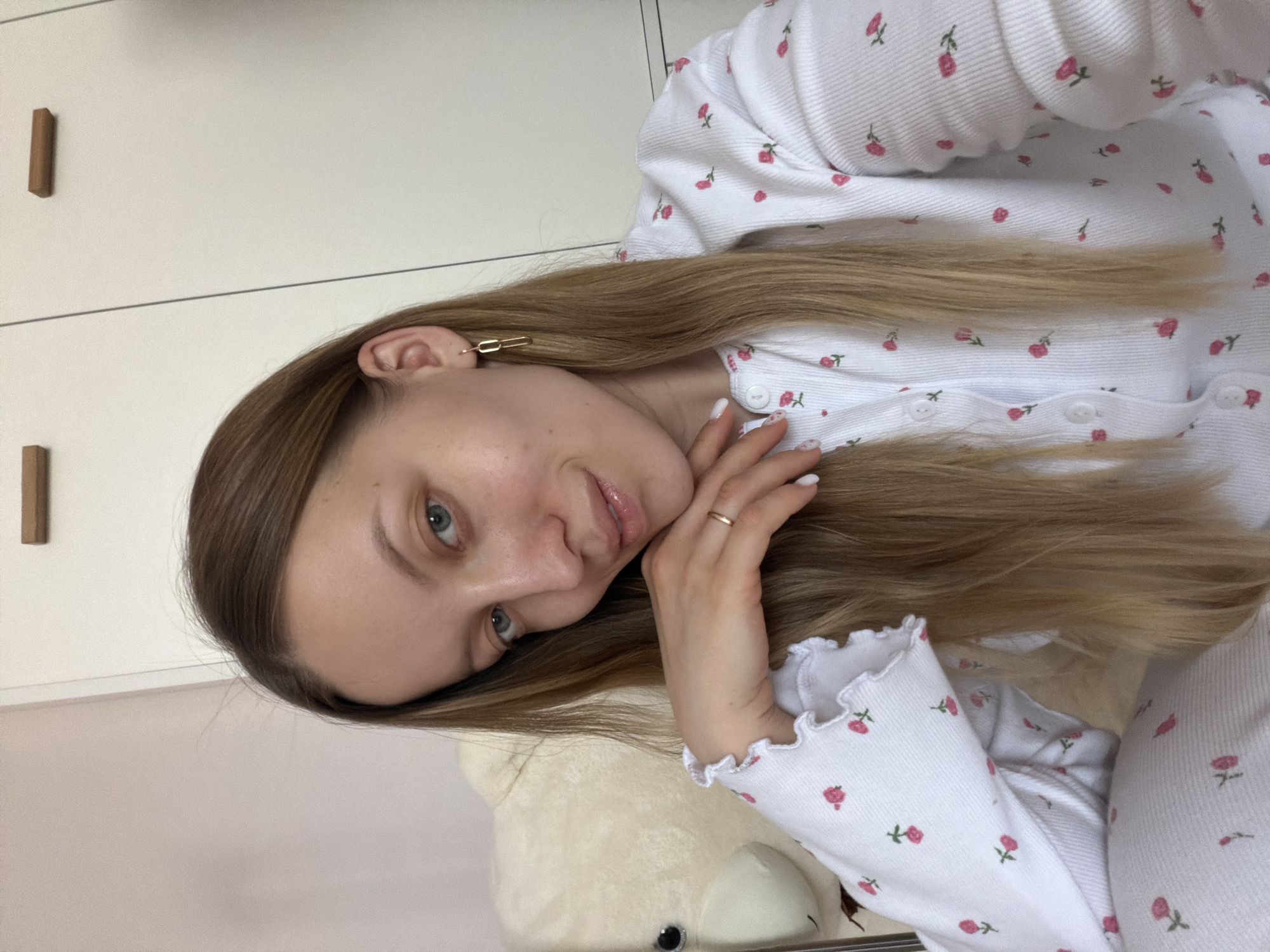 A young woman with long, light brown hair is lying down, resting her cheek on her hand. She is wearing a white long-sleeved top with a small rose print. A gold ring is visible on her finger. She has blue eyes and fair skin. A gold oblong earring is in her left ear. Behind her is a white teddy bear and white furniture with brown handles. The location is likely inside a home. There is no discernible evidence in the image that places the subject at a specific location within Reshetnikovo, Russia.