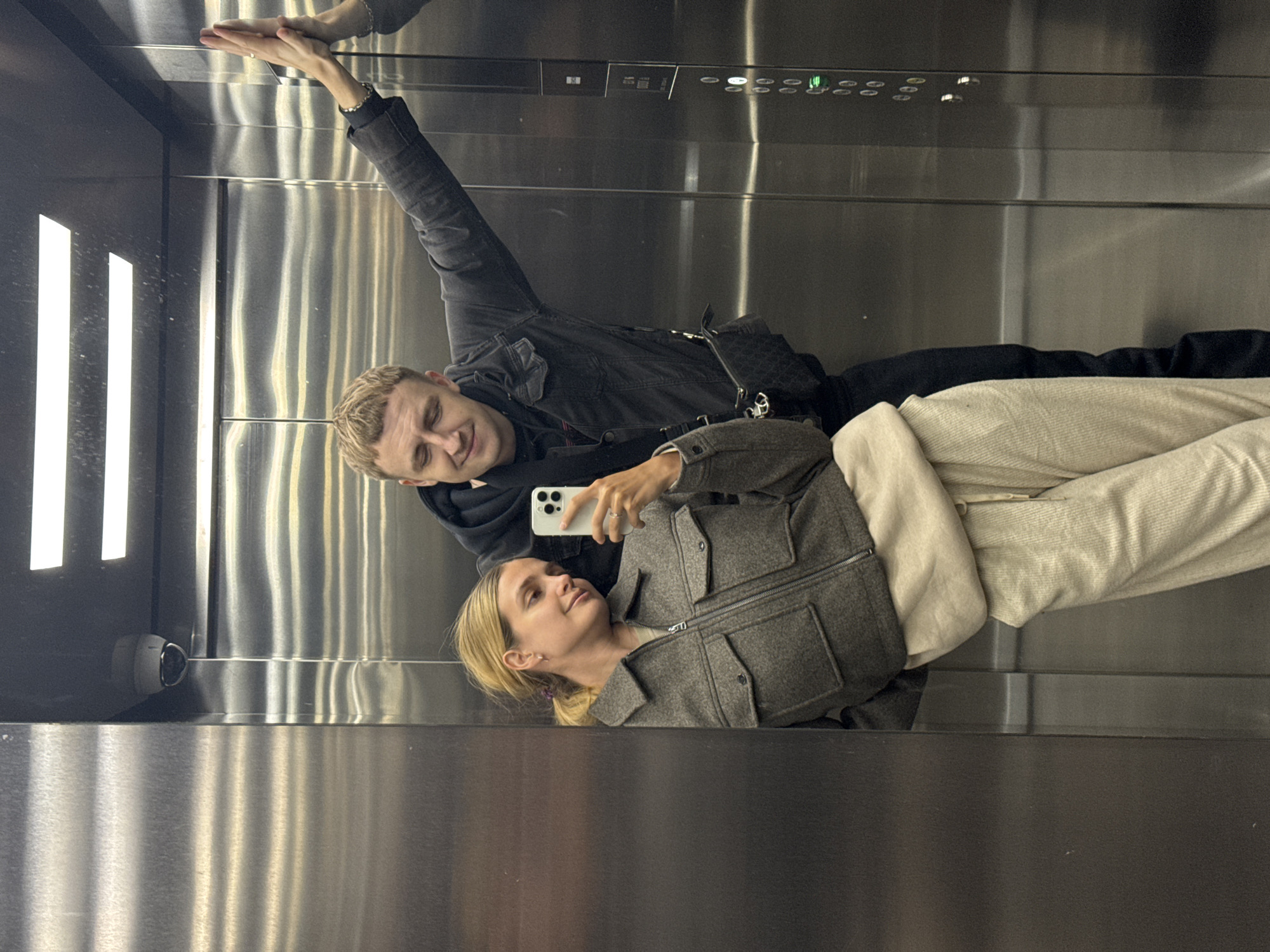 Two individuals are in an elevator, as suggested by the metal interior, the buttons, and the strip lighting. One individual, a male with blond hair, is taking a picture with a smartphone, holding it with his right hand. He is wearing a black jacket. A second individual, a blonde female, is next to him, wearing a gray jacket. The male has one arm raised towards the ceiling. The elevator walls are stainless steel. A security camera is visible in the lower left corner. The city is unknown.
