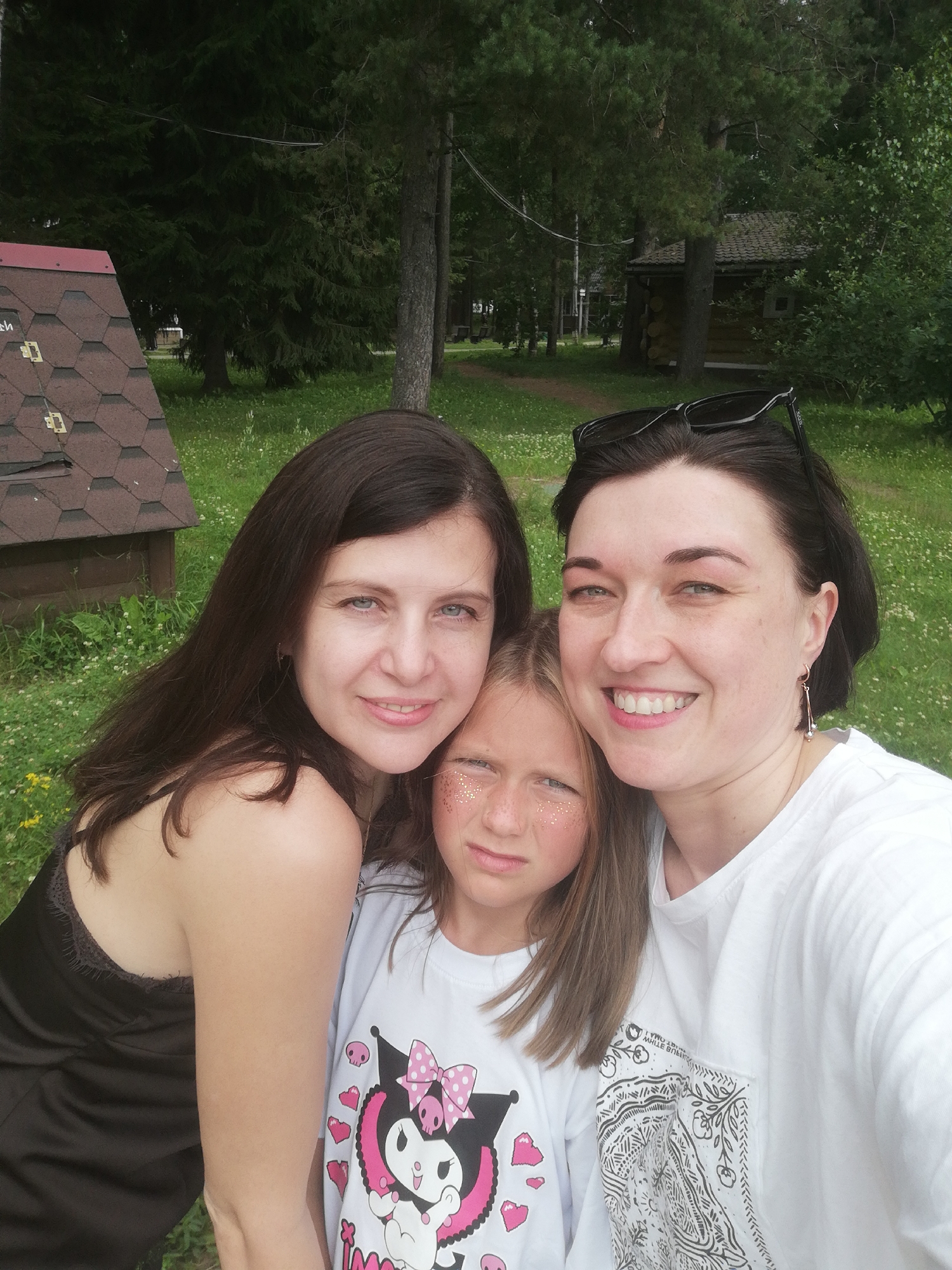 Three females are posing for a selfie outdoors. The person on the left has long dark hair and is wearing a black camisole. The person in the middle is a young girl with blonde hair and is wearing a white t-shirt with a cartoon character on it. She has glitter on her cheeks. The person on the right has dark hair and is wearing a white t-shirt. She has sunglasses perched on top of her head. They are in front of a grassy area with trees and a small wooden structure in the background. The overall setting appears to be a park or recreational area.