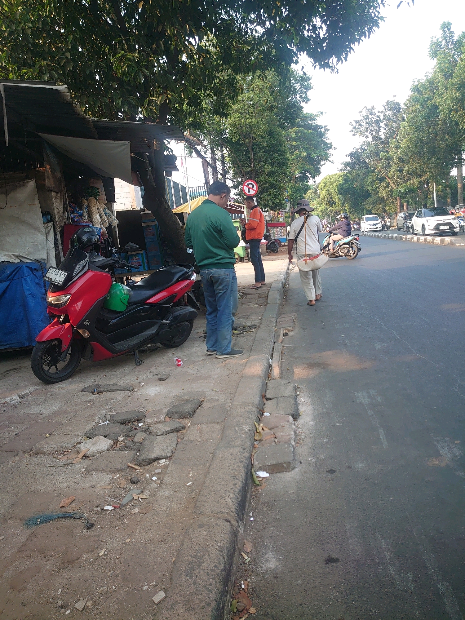 This image depicts a street scene in South Jakarta, Indonesia, likely during daylight. A red and black scooter is parked on the side of a road with a sidewalk. In the foreground, a man in a green jacket and blue jeans stands near the scooter, facing away from the camera. Further down the sidewalk, two individuals are walking away from the camera. One is wearing a light-colored shirt and pants with a straw hat and carrying a bag over their shoulder. The other person is wearing an orange jacket and dark pants. The street is lined with trees, and there are several cars and a motorcycle driving on the road in the background. A small, makeshift stall with items displayed is visible on the left side of the image. The sidewalk appears to be made of concrete and is in disrepair in some sections. There is some litter on the sidewalk and the road. The overall environment suggests a busy urban setting.