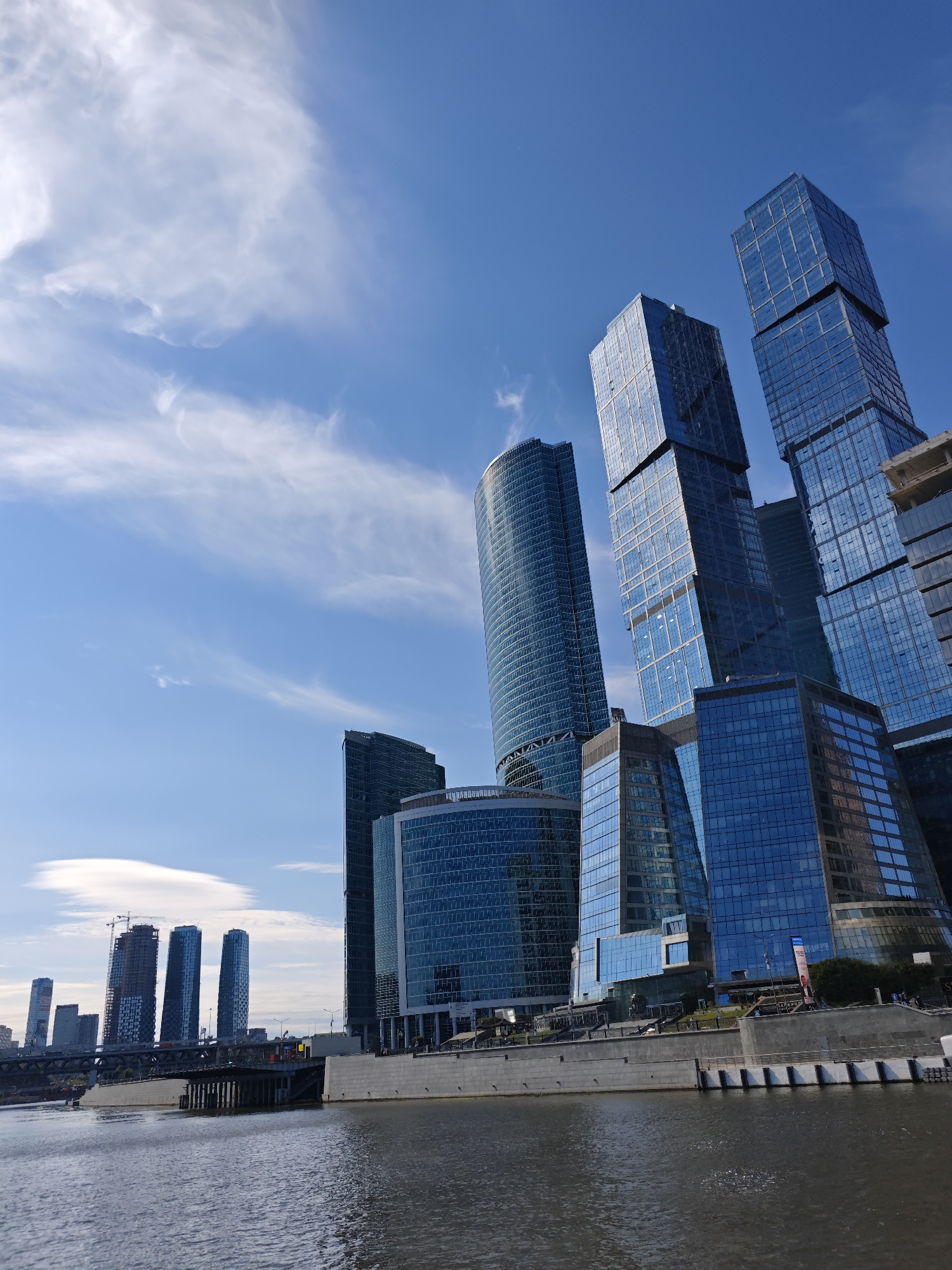 A low-angle, wide shot captures a modern cityscape in Moscow, Russia, dominated by towering glass skyscrapers reflecting a bright blue sky with wispy white clouds. The perspective emphasizes the scale and architectural grandeur of the buildings. In the foreground, a body of water, likely the Moskva River, stretches across the bottom of the frame, its surface rippling gently. A concrete embankment lines the water's edge, with a bridge and some construction cranes visible in the distance, suggesting ongoing development. The skyscrapers, a collection of sleek, geometric structures with dark blue glass facades, are clustered together, creating a dramatic vertical panorama. Their reflective surfaces mirror the sky and surrounding structures, adding depth and dynamism to the scene. The overall impression is one of a bustling, contemporary urban environment with a focus on modern architecture and infrastructure. No individuals are clearly discernible in the image.