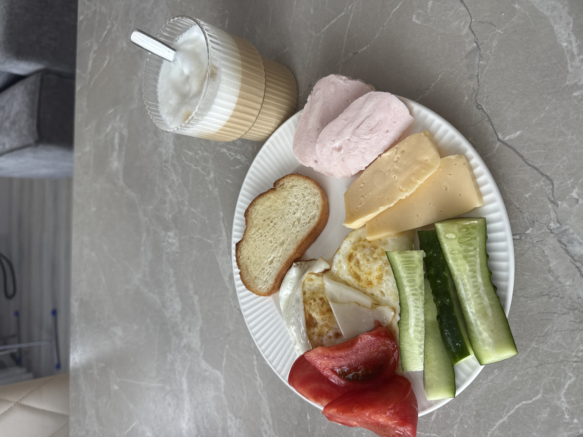 A close-up, eye-level shot captures a breakfast spread on a gray marble countertop. The plate, made of white ceramic with a ribbed texture, holds a variety of food items. In the upper left quadrant of the plate are sliced red tomatoes and two fried eggs, one of which appears to have a slightly runny yolk. To the right of the eggs is a slice of toasted bread. The bottom half of the plate displays sliced cucumbers and several slices of yellow cheese. In the upper right portion of the image, a tall, fluted glass contains a layered beverage, likely coffee with milk or a latte, topped with foam. A silver spoon rests in the drink. In the blurred background, furniture, possibly couches, and a tiled floor are visible, suggesting an indoor setting, likely a home. The image does not contain any information indicating its location in Murom, Russia.