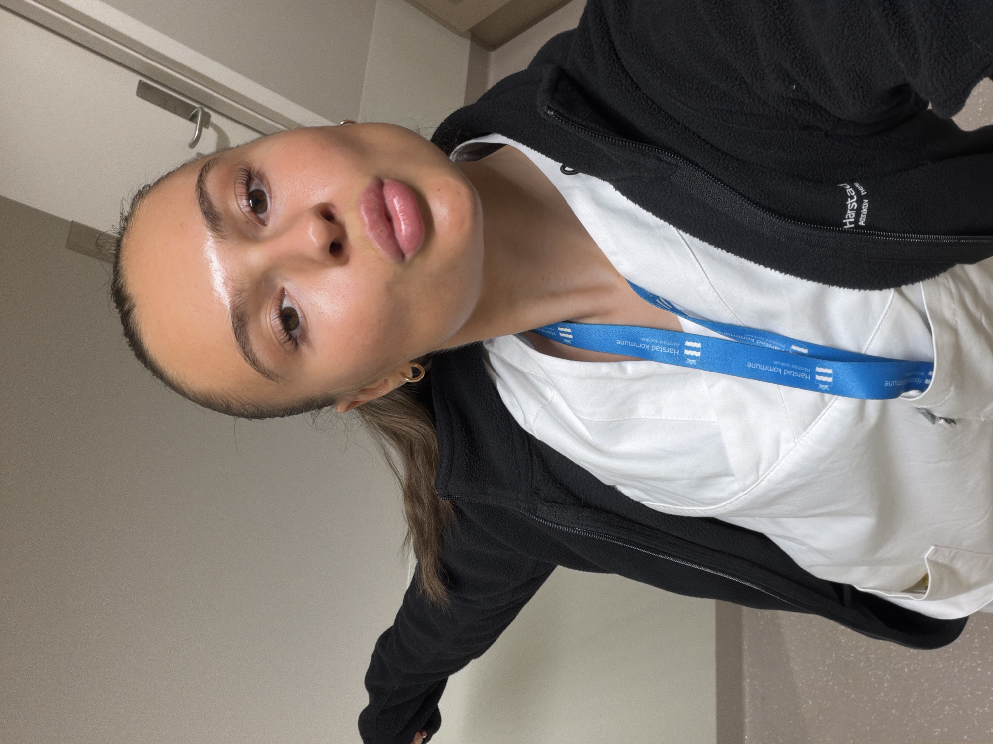 A young woman with her hair in a ponytail, wearing a black zip-up fleece over a white uniform and a blue lanyard with the Harstad kommune logo, poses for a selfie. She is puckering her lips, and her head is turned slightly to the side. She is wearing gold hoop earrings. The background appears to be an indoor setting, possibly a hallway or an office, with a door and cabinetry visible. The floor has a speckled pattern. The lighting suggests it is daytime, but the exact time of day is not discernible. The presence of the Harstad kommune logo on her lanyard indicates that this photo was taken in Harstad, Norway, and that she is likely an employee of the municipality.