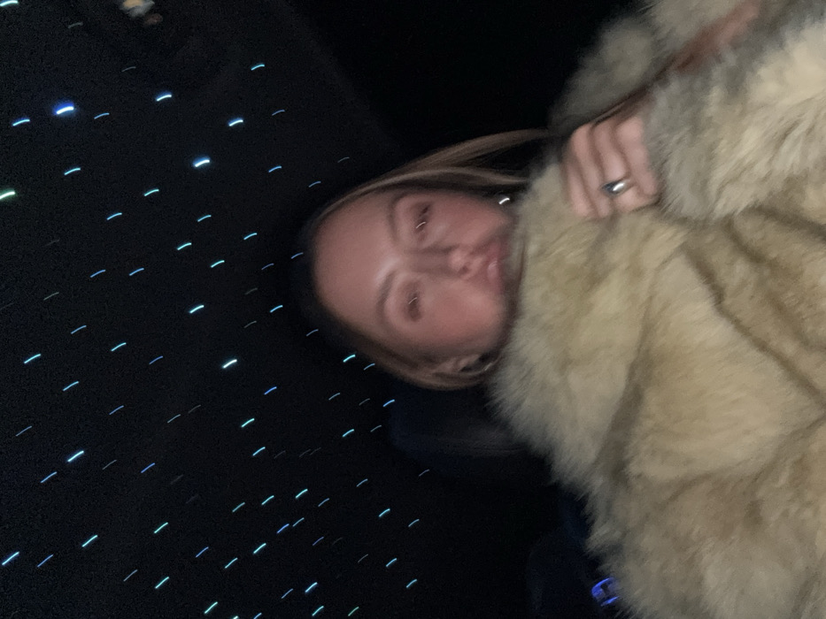 A person with blonde hair is partially visible, face-forward, wearing a large, tan fur coat. A silver ring is on their finger. The background is dark with many small, linear blue lights that resemble stars. The photo is slightly blurry and taken at an angle. The person is positioned to the right of the frame, and the dark, starry background fills the left.