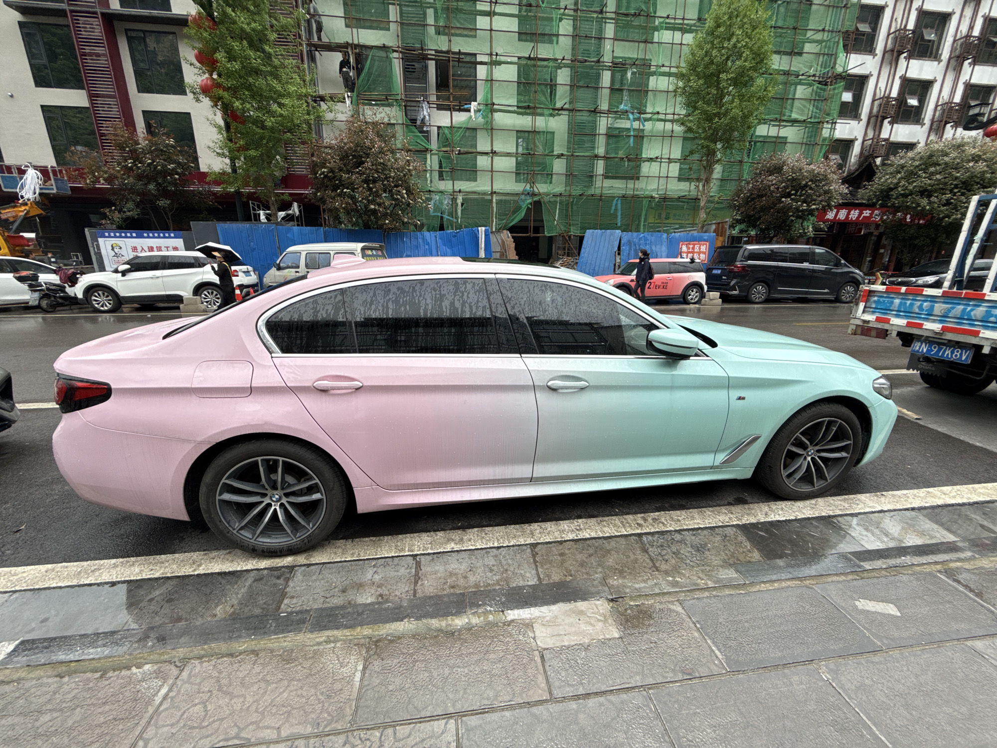 This media file captures a street scene in what appears to be an urban area, likely Wulingyuan, China, given the context. The dominant subject is a BMW sedan, distinctly painted or wrapped in an ombre style, transitioning from light pink at the rear to mint green at the front. The car is wet, suggesting recent rain or damp conditions. Its wheels are dark grey multi-spoke rims.
The pavement is also wet, reflecting the overcast sky. On the right side, there's a sidewalk with large grey tiles. In the background, there's a mix of buildings. Several buildings are under construction or renovation, covered in green scaffolding and blue construction barriers. Trees are scattered throughout the scene.
Other vehicles are visible: a white SUV with a person holding a black umbrella nearby, a black minivan further back, and a red or pink compact car (possibly a Mini EVOKE) to the left of the minivan. Part of a blue and white truck with a wooden bed is visible on the far right.
Visible text from signs includes:
- "进入建筑工地" (Jìnrù jiànzhú gōngdì) meaning "Enter construction site."
- "施工区域" (Shīgōng qūyù) meaning "Construction area."
- "防坠物" (Fáng zhuì wù) meaning "Beware of falling objects" or "Anti-falling objects."
- "湖南特产购对" (Húnán tèchǎn gòu duì) possibly related to Hunan local specialties shopping.
- A license plate "豫N-97K8V".
- "2 EV.KE" on the red/pink compact car.
The overall impression is a bustling urban environment on a damp, possibly rainy day.