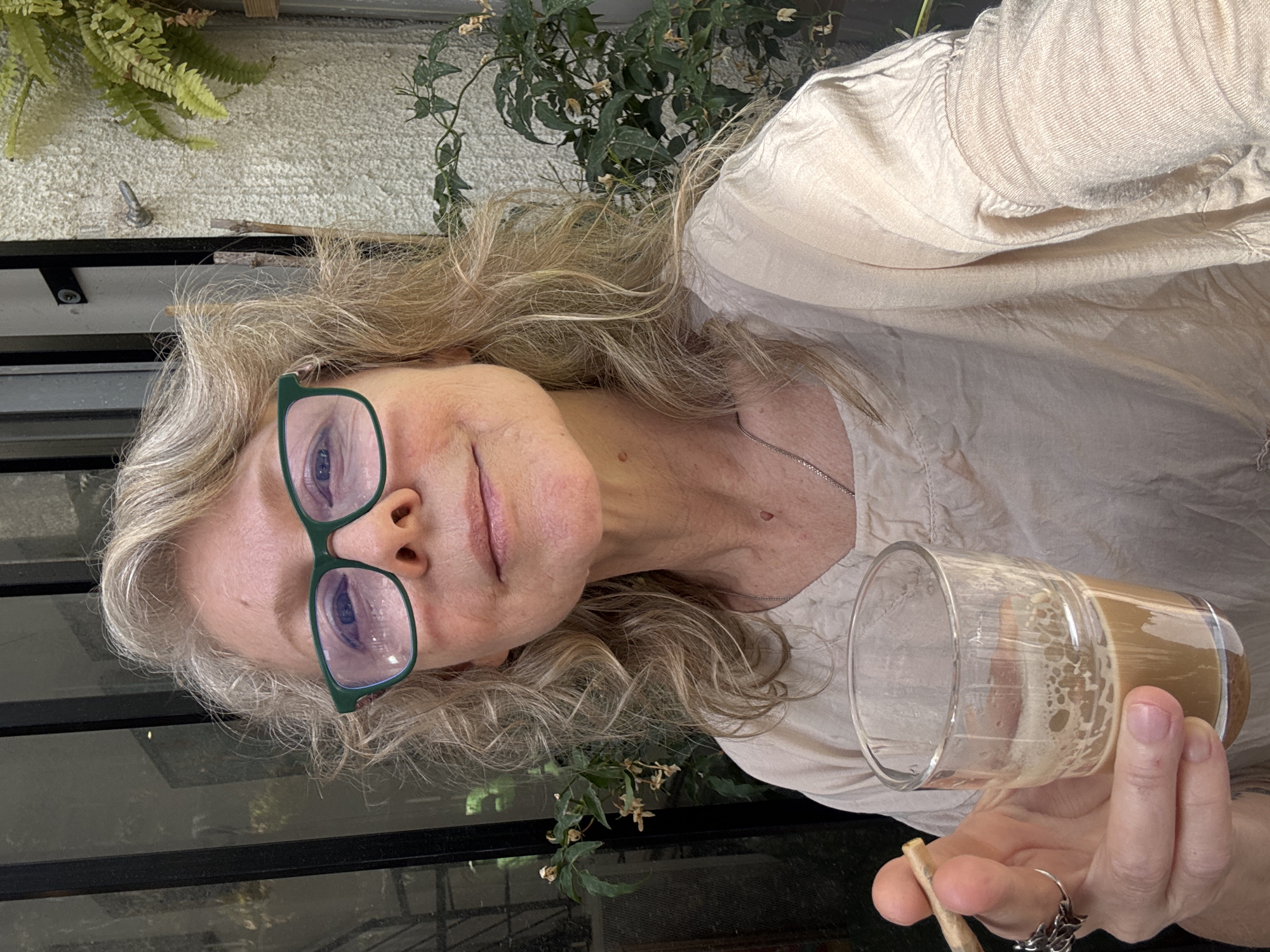 A middle-aged woman with blonde curly hair and green glasses is holding a partially empty glass of coffee and a hand-rolled cigarette. She is wearing a beige shirt and a silver necklace. She is in an outdoor setting, likely a patio or balcony. There are plants visible in the background, along with a black railing. The setting appears to be residential and does not explicitly suggest a location in Tel-Aviv, Israel.