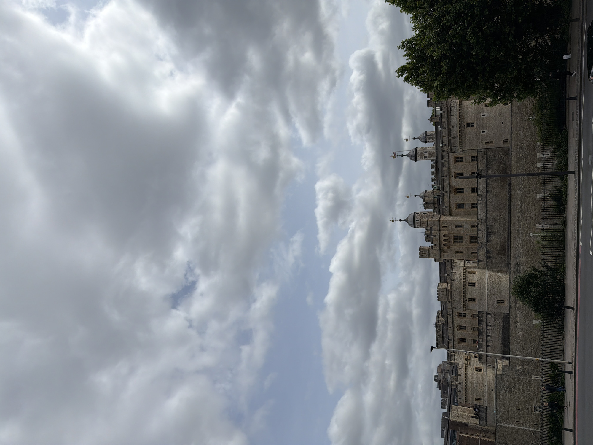 This image depicts the Tower of London, a historic castle located in the City of London, United Kingdom. The imposing stone fortress occupies the middle ground, featuring multiple circular and square towers, crenellated battlements, and visible windows. Several finials or weathervanes are visible atop the various towers. The sky above is largely covered by grey stratocumulus clouds, with intermittent patches of lighter blue sky visible, particularly in the upper left and center-right areas.
In the foreground, a multi-lane road runs horizontally across the bottom of the frame, with a grey pavement bordering it. A black metal fence separates the pavement from a lower-level area immediately adjacent to the Tower's base. A tall, black lamppost stands prominently on the pavement, slightly left of center. To the right, a large, dark green leafy tree partially obscures the lower section of the Tower. Two individuals are discernible on the pavement to the left of the lamppost; one is standing, and another appears to be walking. The overall light suggests an overcast day.